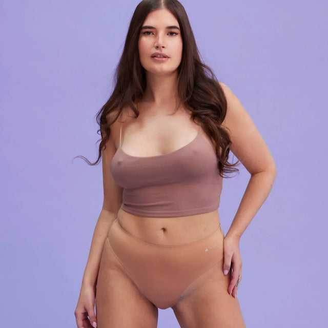 Eine Frau mit langen braunen Haaren kniet vor einem hellvioletten Hintergrund auf dem Boden. Sie trägt ein lila Crop-Top und nackte Unterwäsche mit hoher Taille.