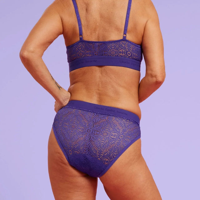 Eine Frau mit langen grauen Haaren, von hinten gesehen, trägt ein passendes Set aus lila Spitzenunterwäsche und einem Bralette. Sie steht vor einem pastelllila Hintergrund und zeigt eine Tätowierung auf ihrem oberen Rücken.