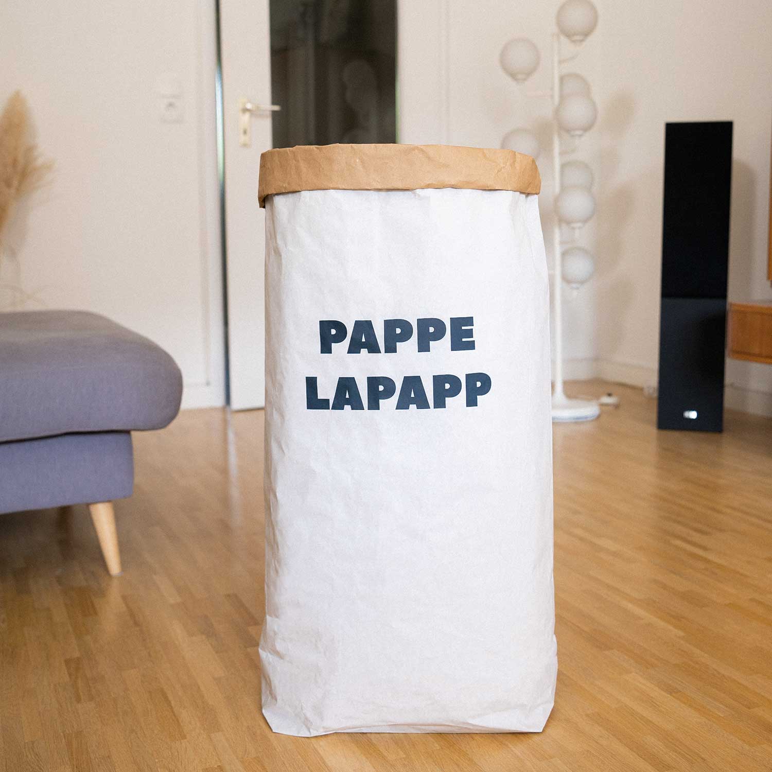 Ein großer weißer Papiersack mit der Aufschrift PAPPE LAPAPP steht auf einem Holzboden in einem modernen Wohnzimmer mit einem grauen Sofa und einer dekorativen Lampe im Hintergrund.