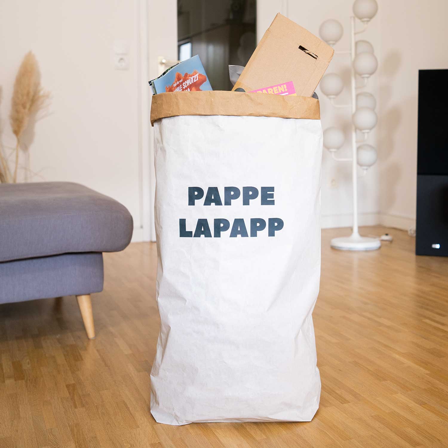 Eine große weiße Papiertüte mit der Aufschrift PAPPE LAPAPP steht auf einem Holzboden, gefüllt mit Pappartikeln. Die Umgebung scheint ein Wohnzimmer zu sein, mit einem grauen Sofa, einer Stehlampe und dekorativen Gegenständen im Hintergrund.