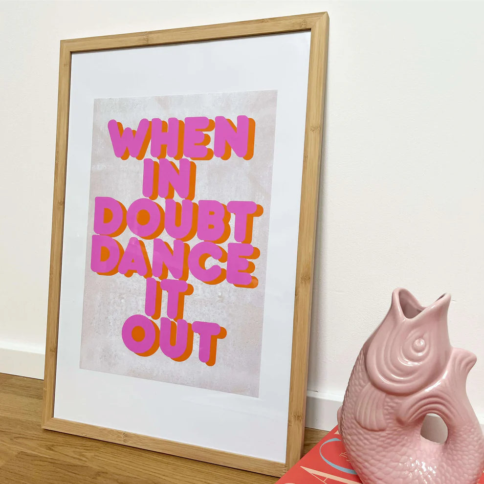 Ein gerahmter Druck mit dem fetten rosa-orangen Text WHEN IN DOUBT DANCE IT OUT steht auf dem Boden neben einer rosa Keramikfischvase und einigen gestapelten Büchern.