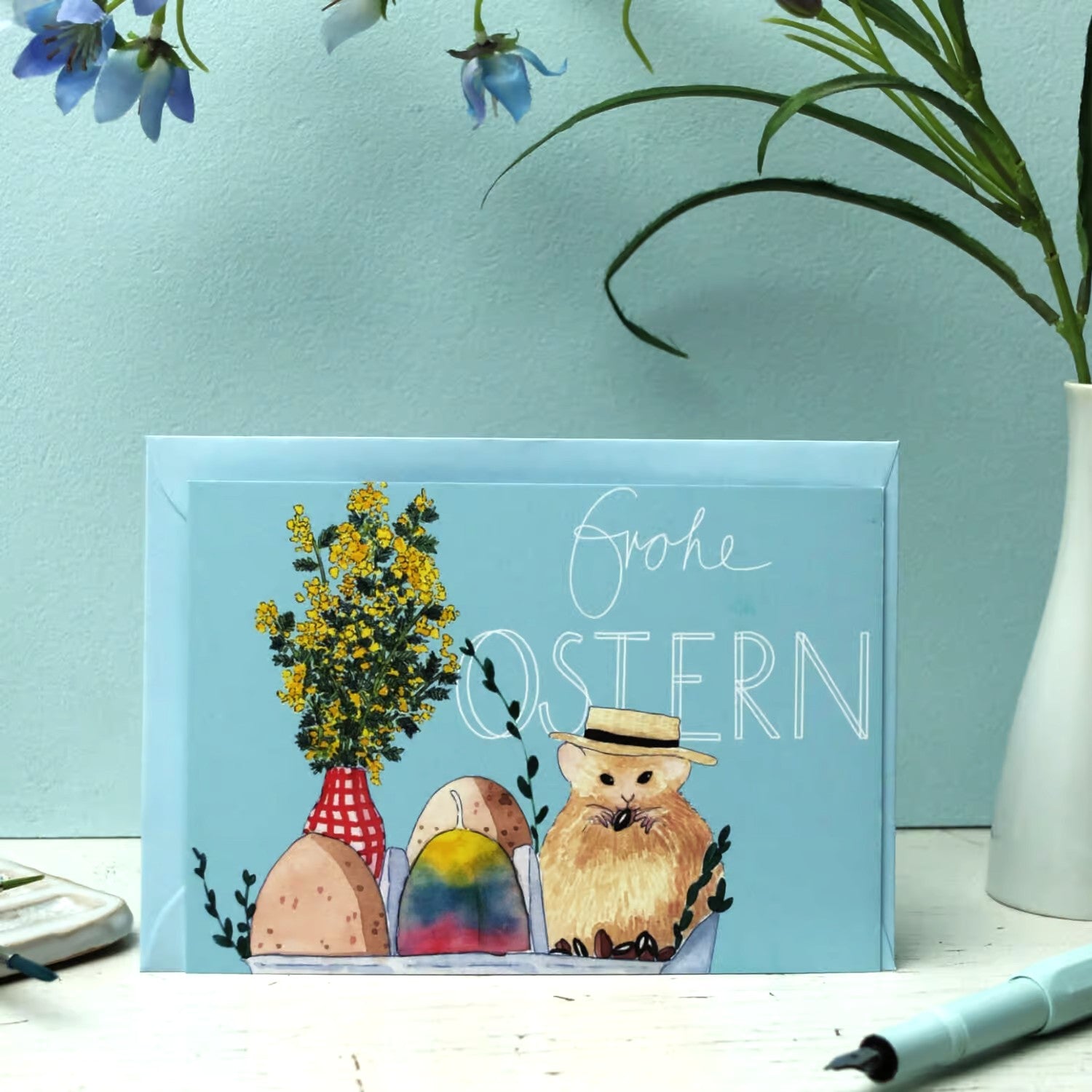 Eine Grußkarte mit Frohe Ostern und einer Illustration einer Katze mit Hut, bunten Ostereiern und gelben Blumen in einer Vase vor einem hellblauen Hintergrund mit Pflanzen in der Nähe.