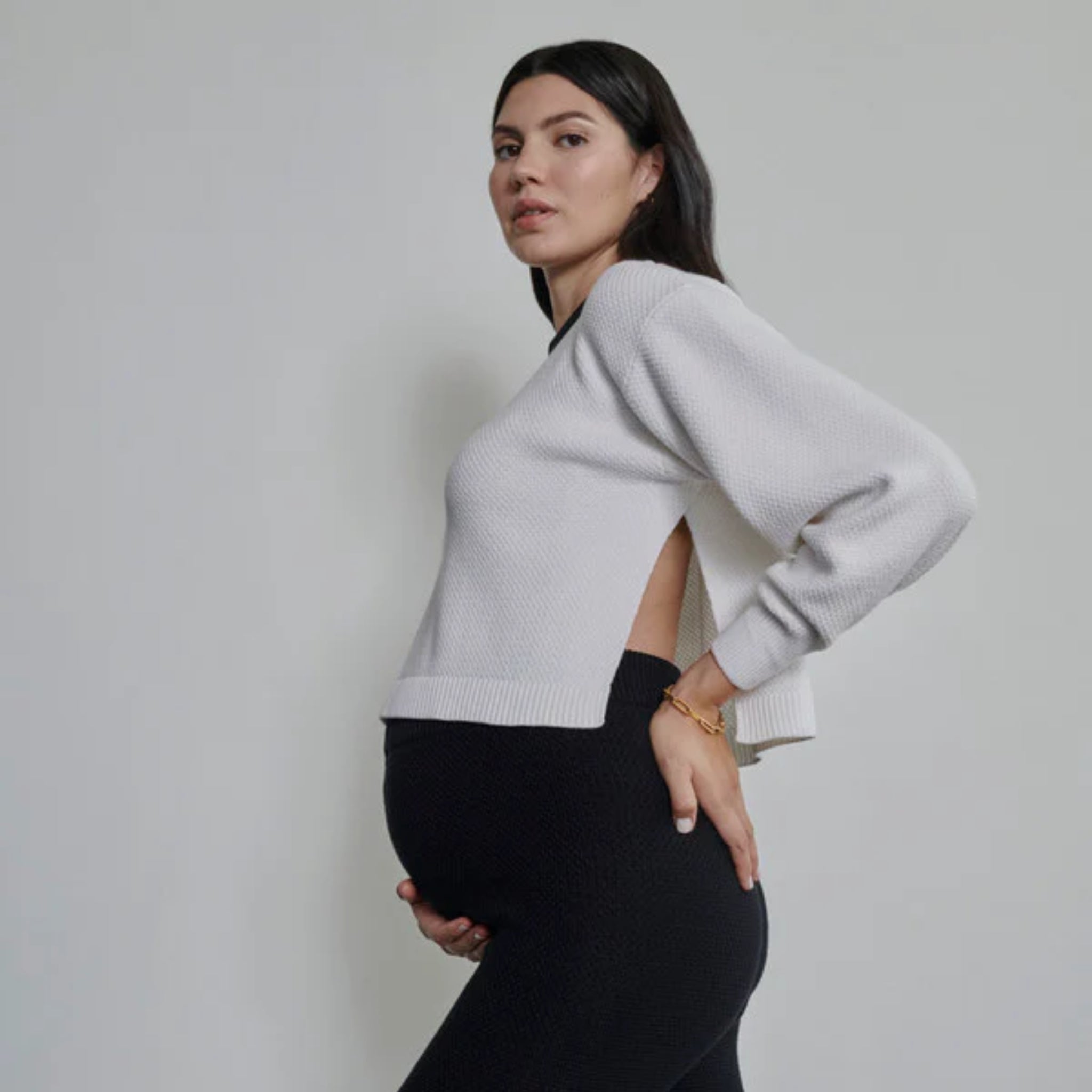 Eine schwangere Person, die schwarze Leggings und einen hellgrauen, offenen Pullover trägt, steht im Profil vor einem schlichten, hellen Hintergrund, hält ihren Bauch und schaut in die Kamera.
