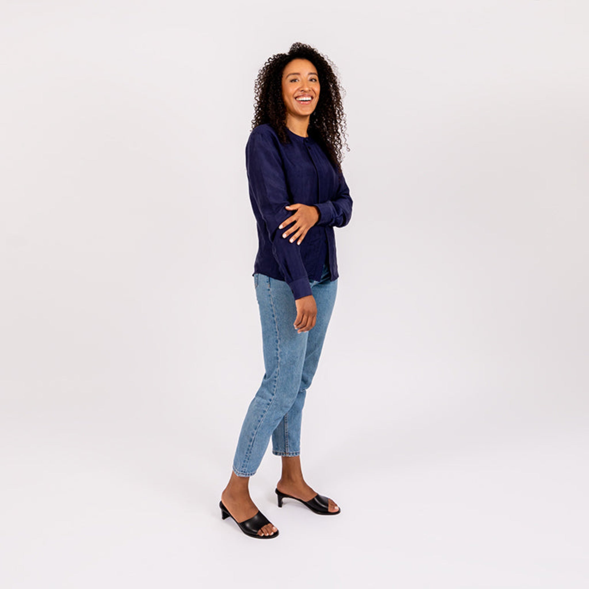 Frau mit lockigem Haar, in blauer Bluse und Jeans, lächelt und steht vor weißem Hintergrund.