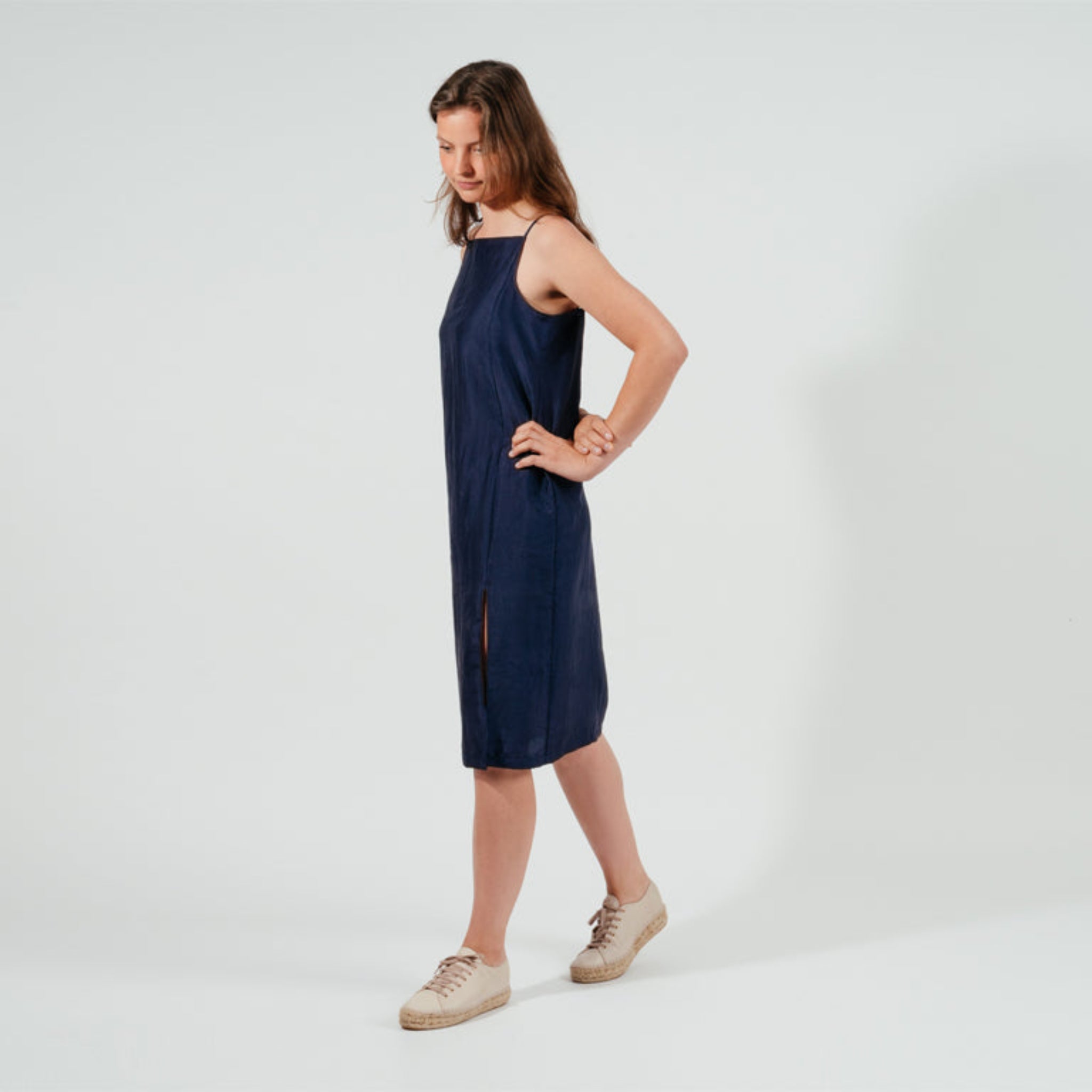 Frau in einem dunkelblauen Kleid und weißen Sneakers steht auf weißem Hintergrund mit Blick nach unten.