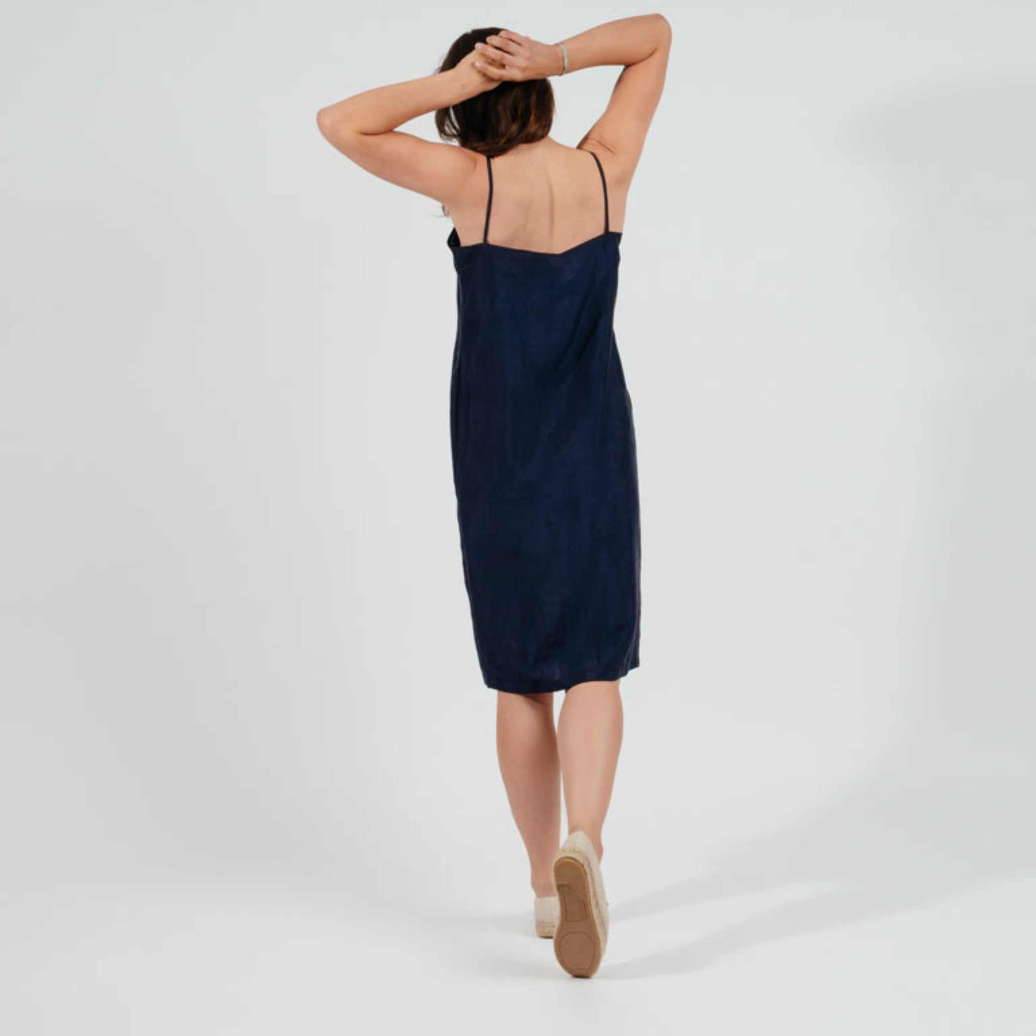 Frau in einem blauen Trägerkleid, mit dem Rücken zur Kamera und den Händen über dem Kopf, geht auf weißem Hintergrund.