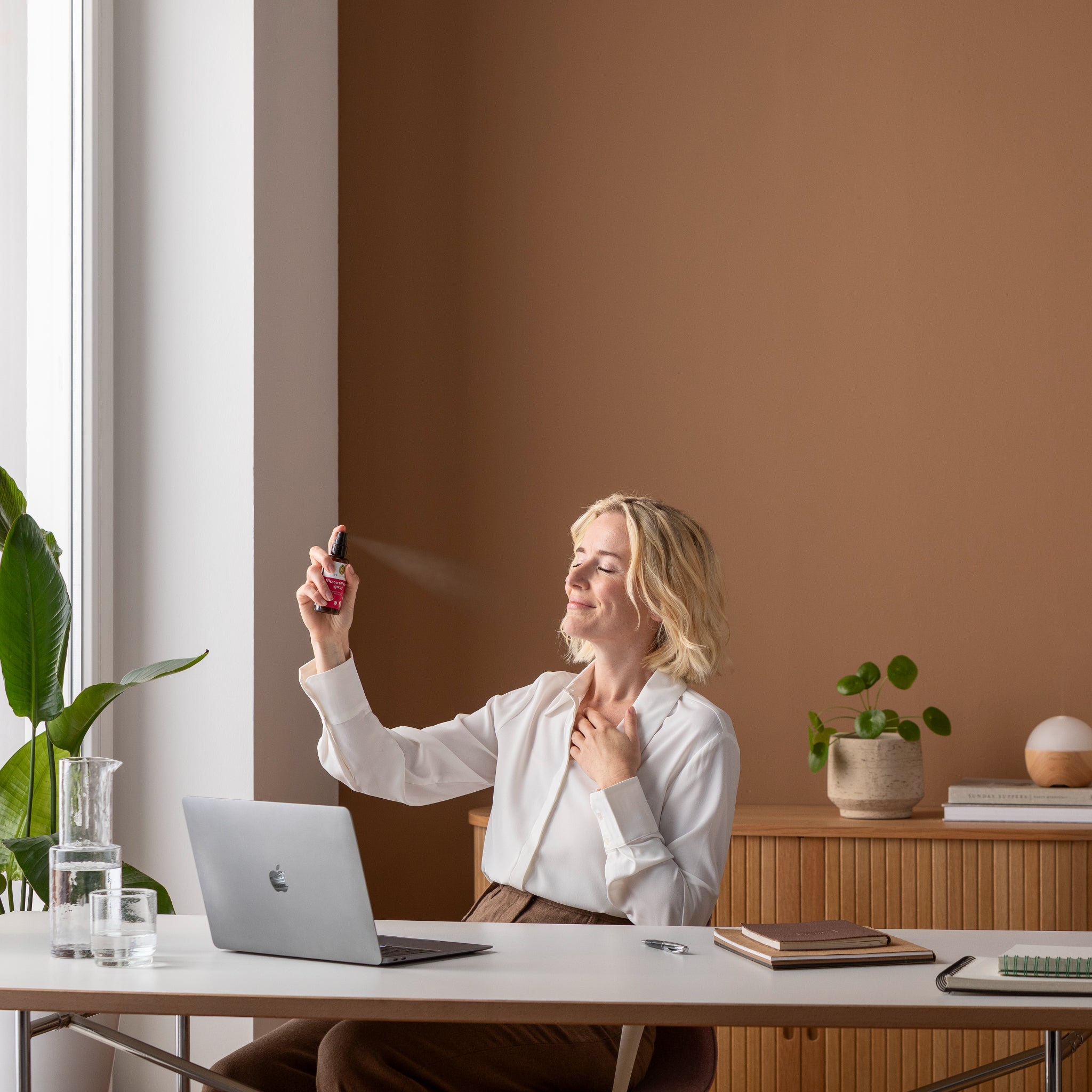 Eine Frau, die an einem Schreibtisch mit einem Laptop sitzt, besprüht ihr Gesicht mit Nebel und lächelt. Auf dem Schreibtisch stehen Notizbücher, ein Glas Wasser und Pflanzen, eine braune Wand und natürliches Licht aus einem nahe gelegenen Fenster.