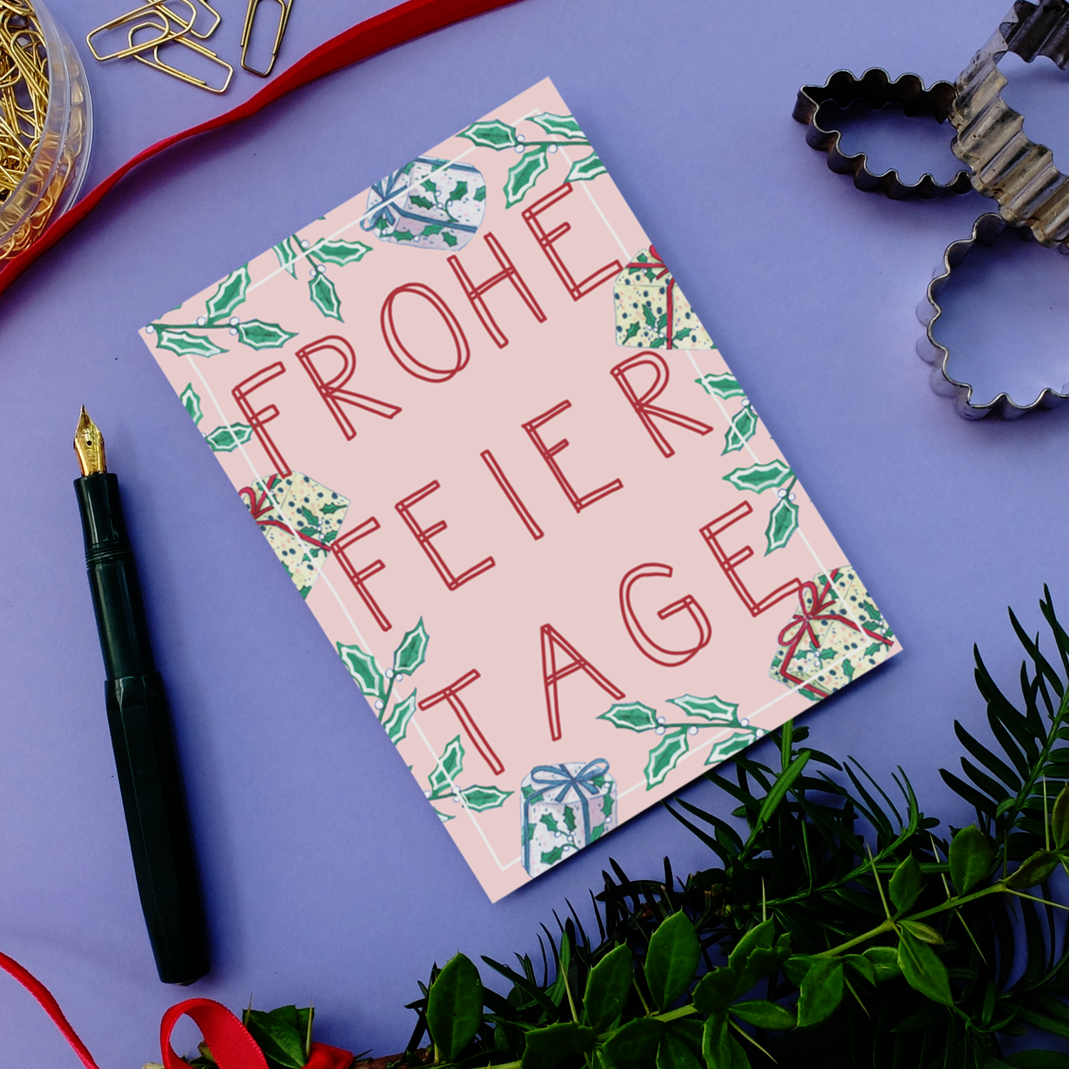 Eine festliche Karte mit der Aufschrift Frohe Feiertage" in roten Buchstaben, verziert mit Stechpalmen und Schleifen, liegt auf einer lila Fläche neben einem Füllfederhalter, Ausstechformen, Büroklammern und grünem Laub.
