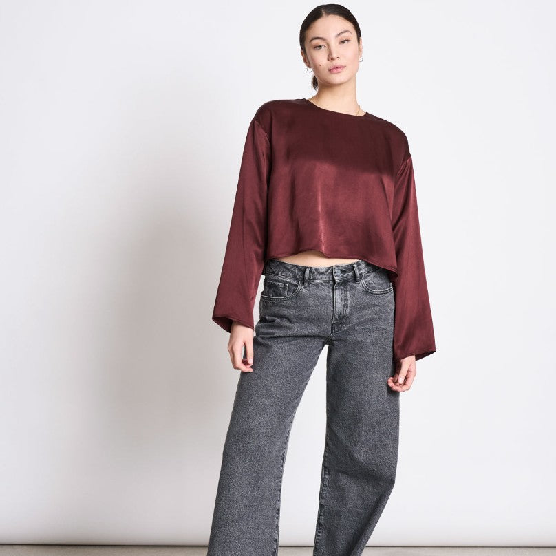 Eine Frau steht vor einem schlichten weißen Hintergrund und trägt ein lockeres, langärmeliges kastanienbraunes Satin-Crop-Top und graue Jeans mit hoher Taille. Sie hat ihr Haar zurückgebunden und posiert mit einer entspannten Hand an ihrer Seite.