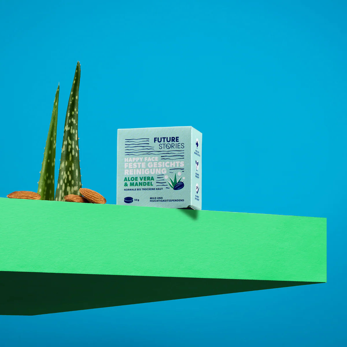 Eine Schachtel Future Stories Gesichtsreinigungsriegel mit Aloe Vera und Mandarine steht auf einem grünen Podest neben zwei Aloe-Vera-Blättern und zwei Mandeln vor einem leuchtend blauen Hintergrund.