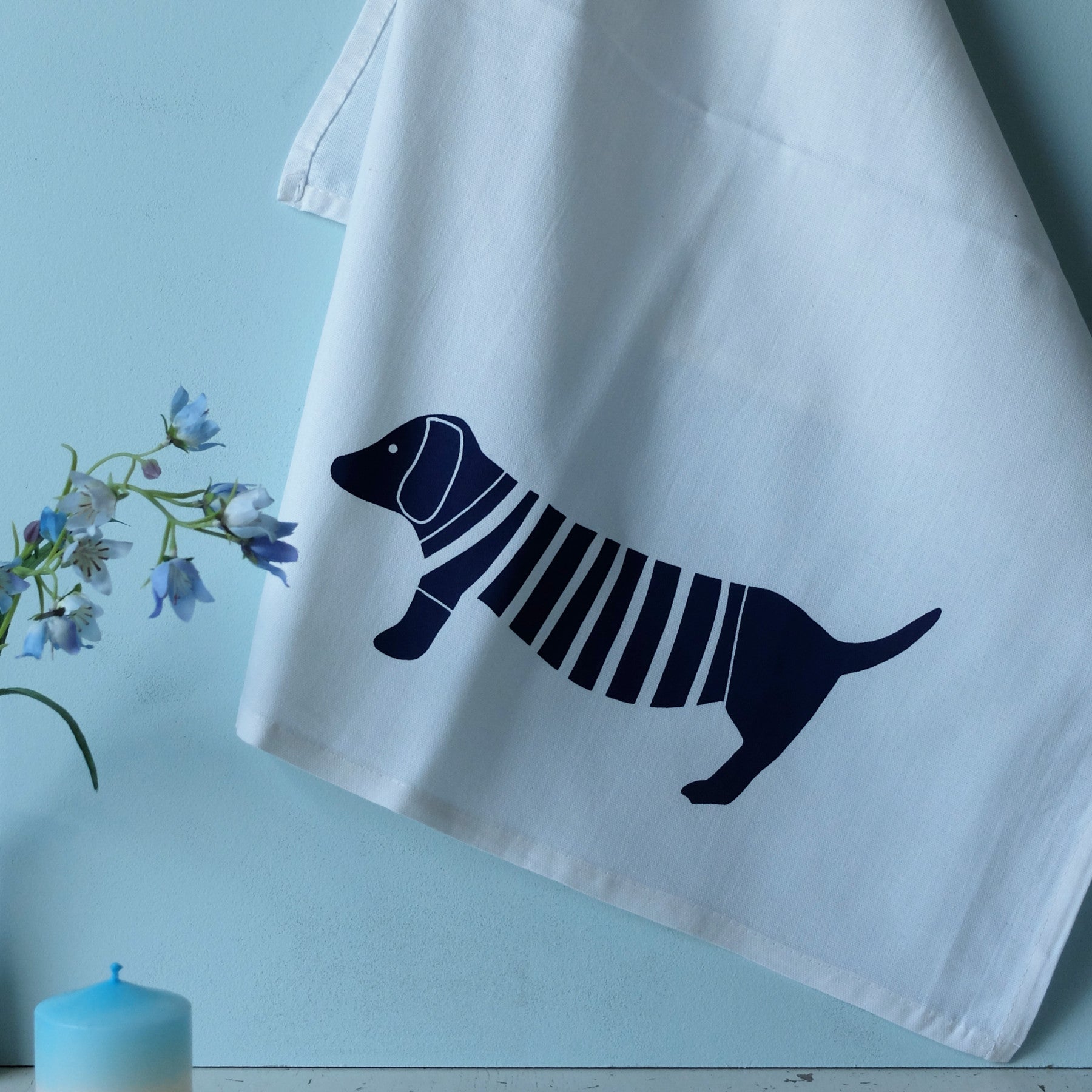 An einer hellblauen Wand hängt ein weißes Tuch mit einer einfachen, marineblauen Illustration eines Dackels, der einen gestreiften Pullover trägt. In der Nähe sind eine kleine blaue Kerze und eine Vase mit lila Blumen zu sehen.