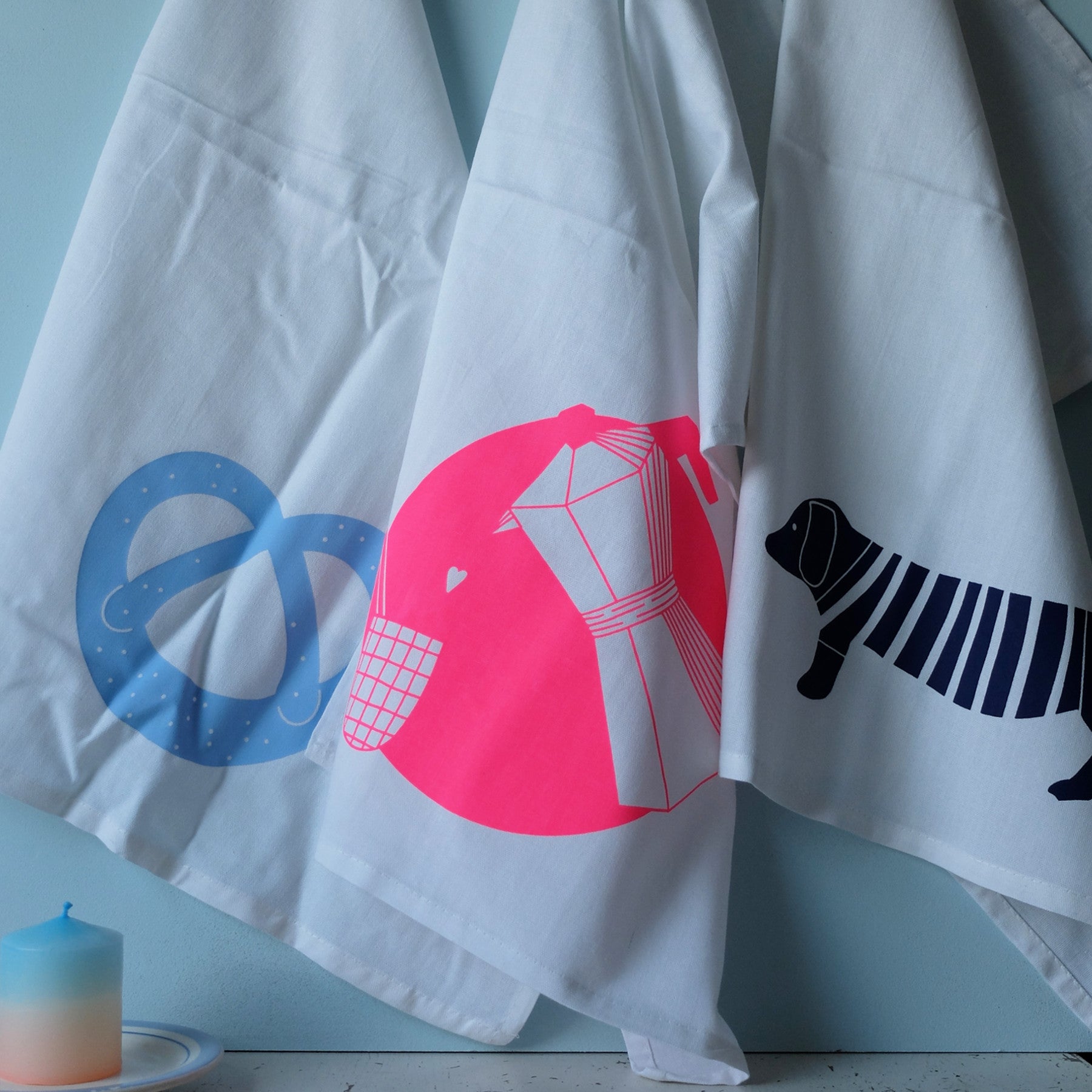 Drei weiße Geschirrtücher hängen nebeneinander, jedes mit einer auffälligen Grafik: eine blaue Brezel, eine rosa Mokkakanne mit Tasse und ein schwarzer Dackel im gestreiften Pullover. Daneben steht eine blau-weiße Kerze.