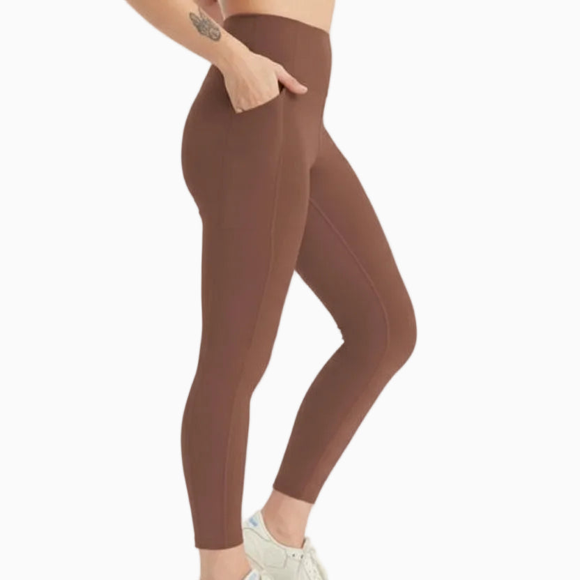 Eine Person in braunen Leggings mit hoher Taille und weißen Turnschuhen steht mit einer Hand in der Tasche vor einem schlichten hellen Hintergrund. 