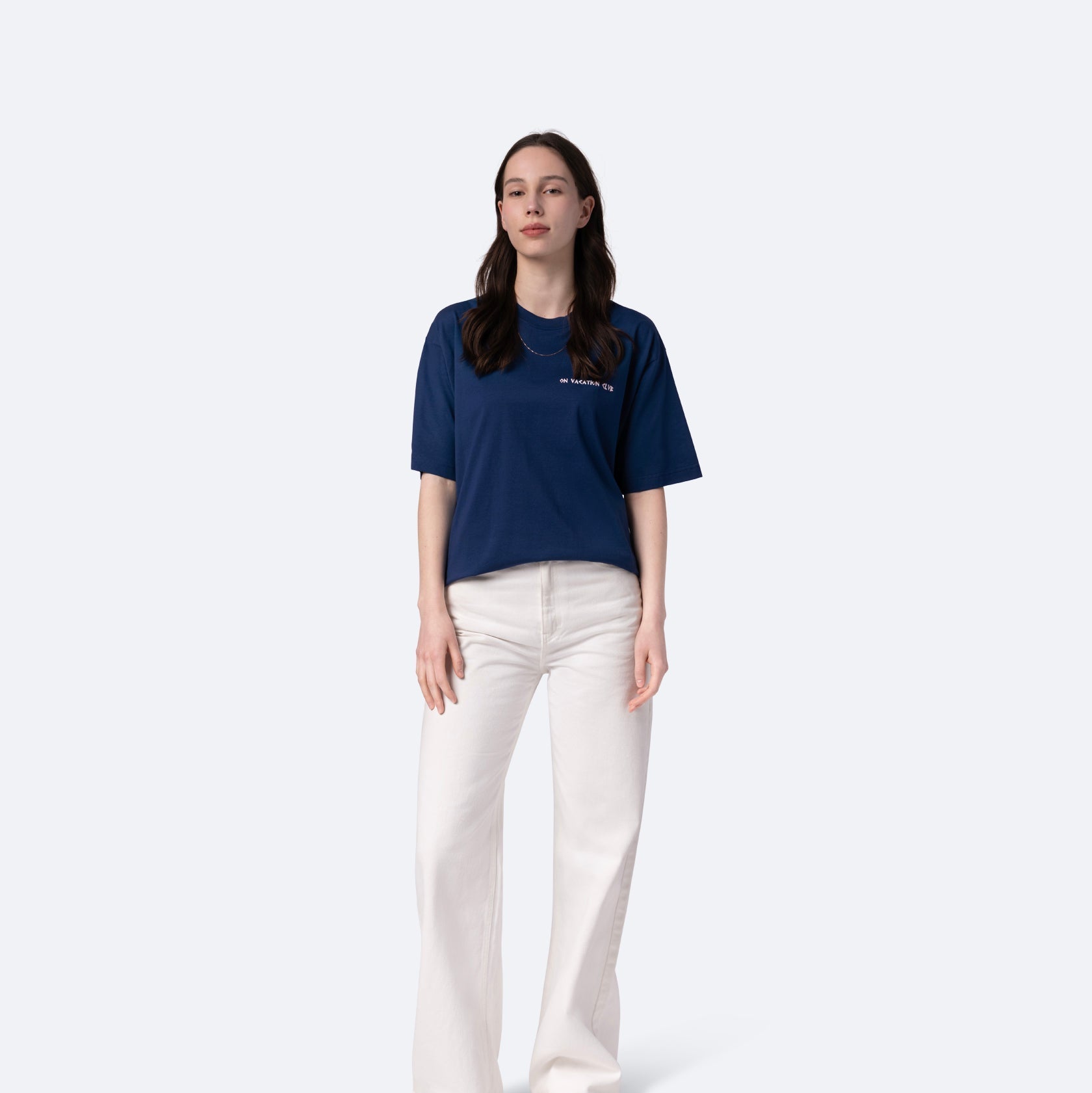 Eine Frau steht vor einem schlichten, hellen Hintergrund. Sie trägt ein marineblaues T-Shirt und eine weiße Hose mit hoher Taille. Sie hat langes braunes Haar und einen neutralen Gesichtsausdruck, eine Hand steckt in ihrer Tasche.