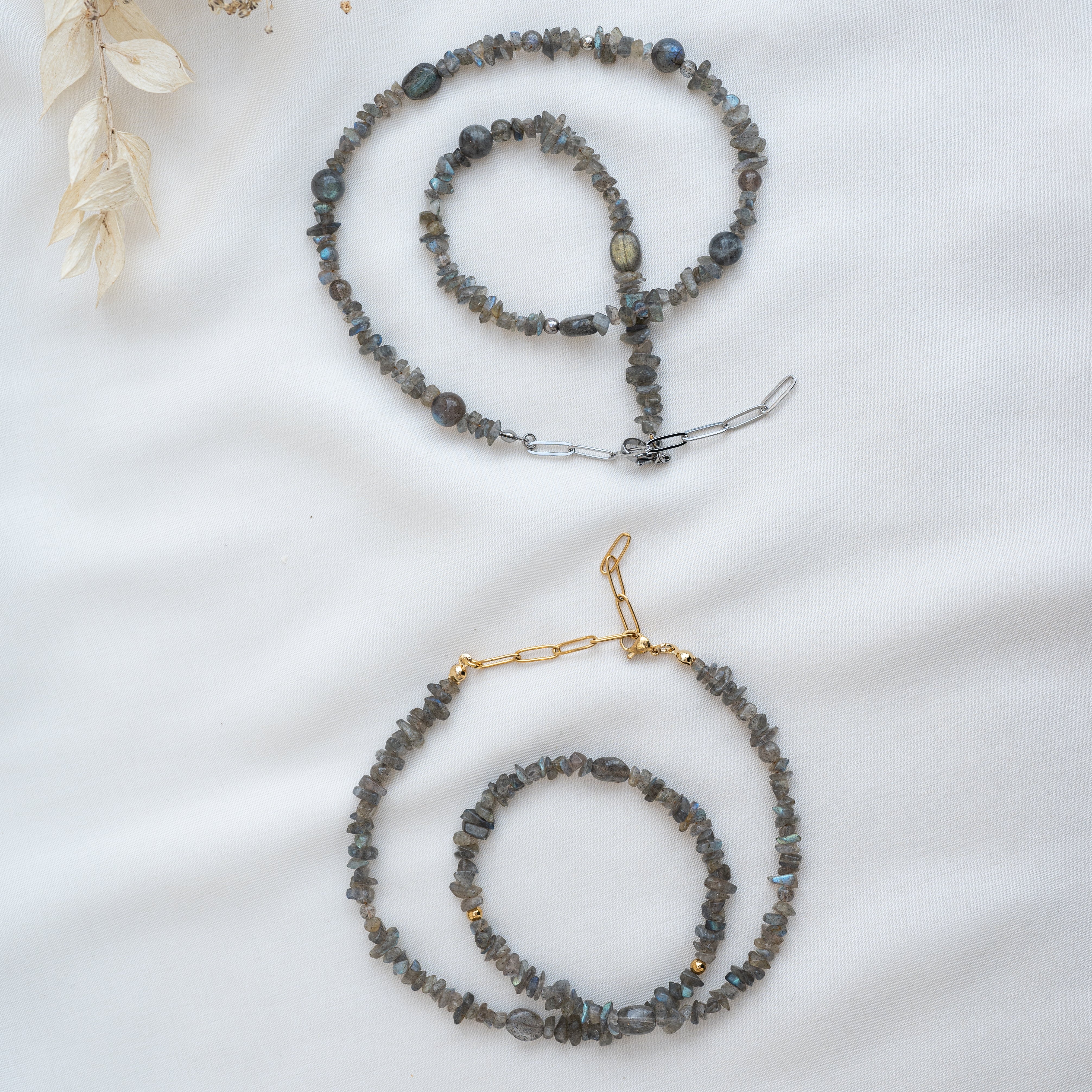 Zwei Perlenketten mit grauen Steinen und goldenen oder silbernen Verschlüssen sind auf einem weißen Stoffhintergrund abgebildet. Getrocknetes beiges Laub ziert die obere linke Ecke des Bildes.