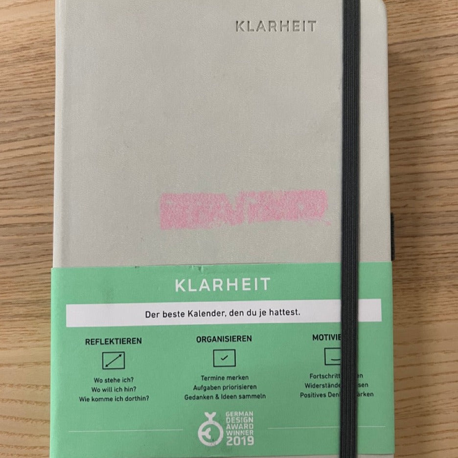 Notizbuch mit der Aufschrift "KLARHEIT" und Gewinner des German Design Award 2019, auf einem Holztisch liegend.