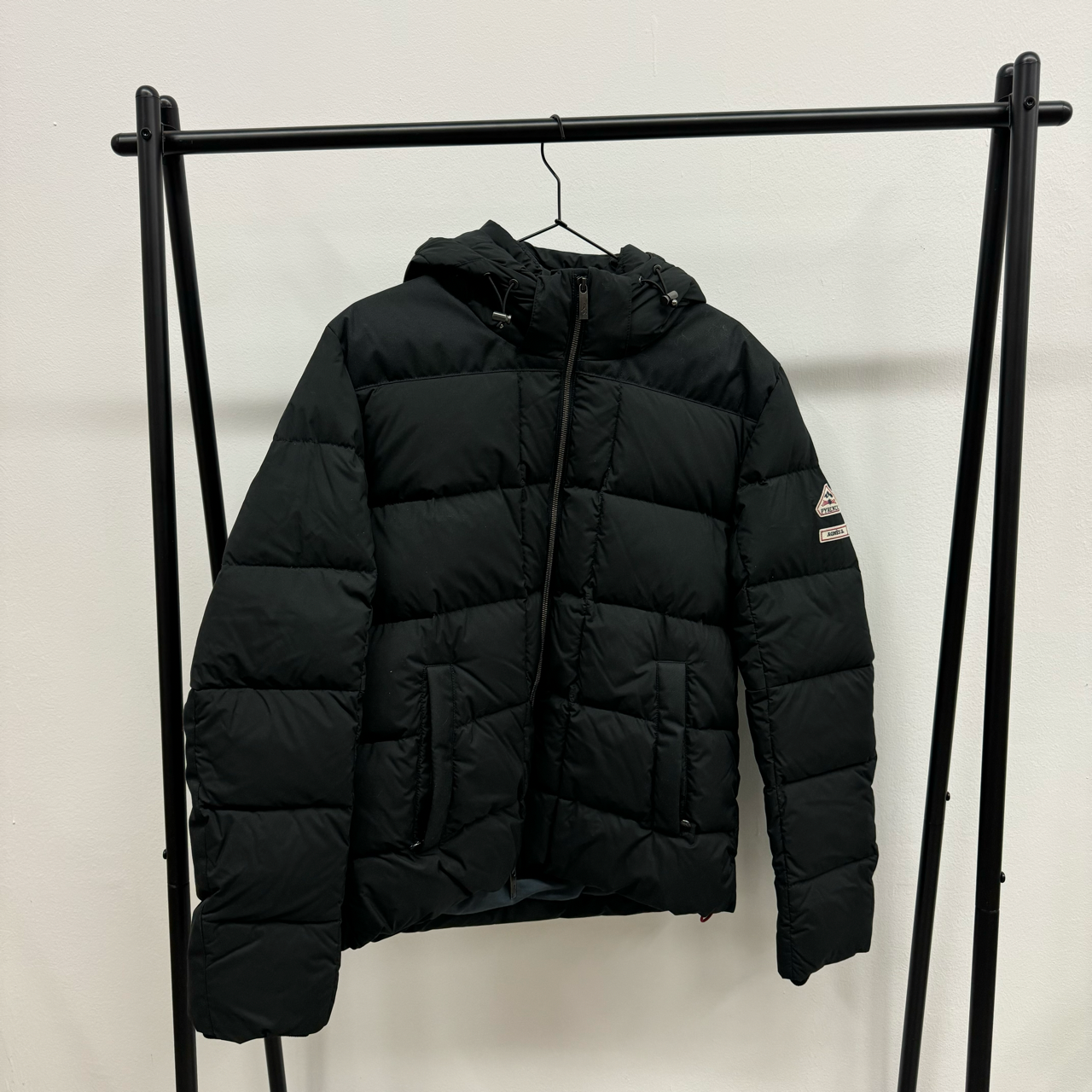 Schwarze, gepolsterte Winterjacke mit Kapuze hängt an einem Kleiderständer vor weißem Hintergrund.