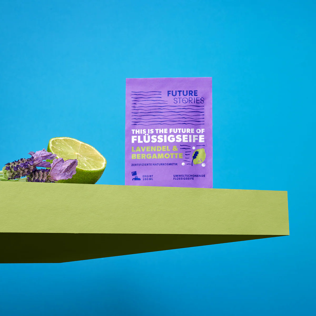 Ein Tütchen Future Stories Lavendel-Bergamotte-Flüssigseife steht aufrecht auf einem grünen Podest, daneben eine halbe Limette und Lavendelzweige vor einem hellblauen Hintergrund.