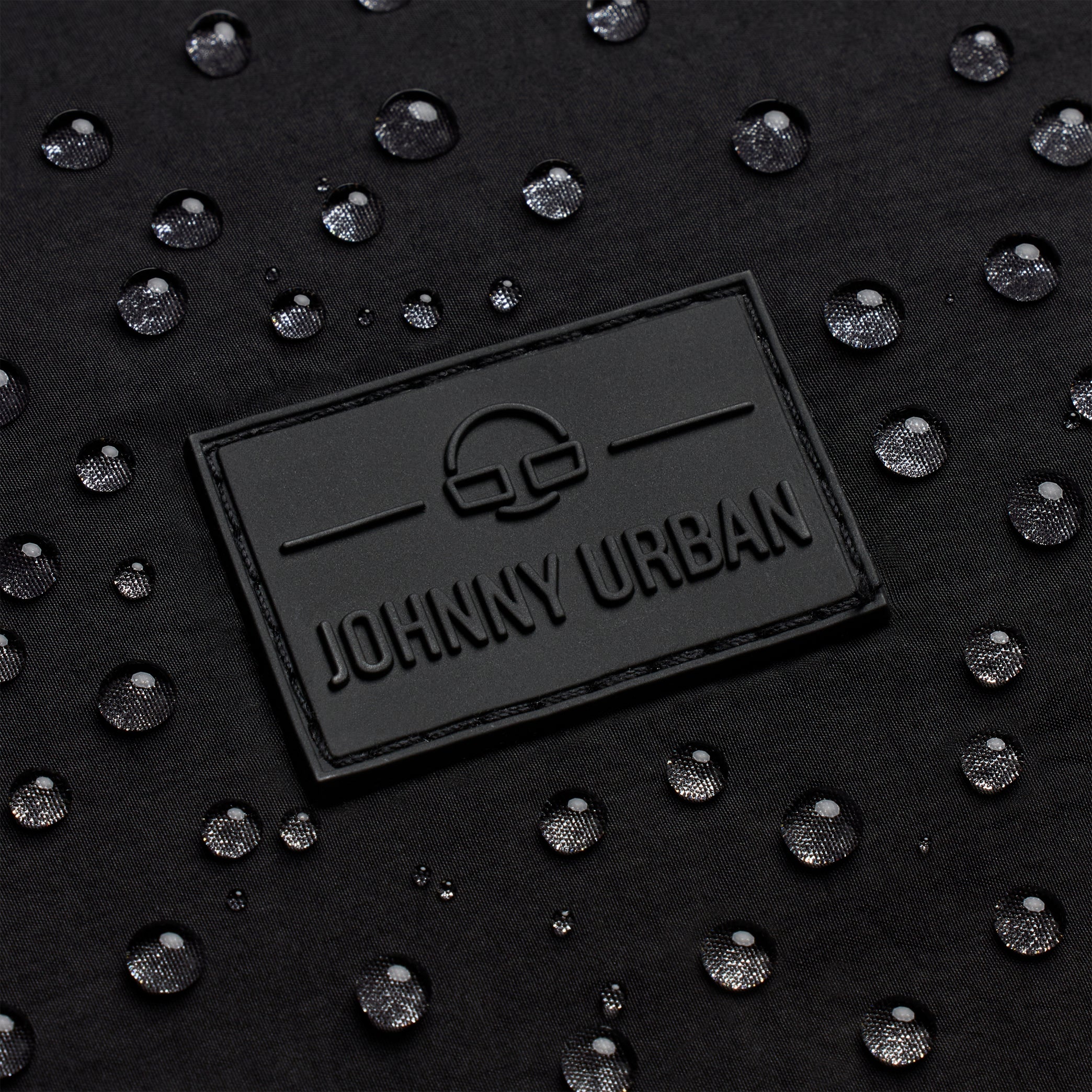Nahaufnahme eines schwarzen Stoffes mit Wassertropfen auf seiner Oberfläche und einem rechteckigen schwarzen Aufnäher mit der Aufschrift JOHNNY URBAN und einem minimalistischen Logo über dem Text.