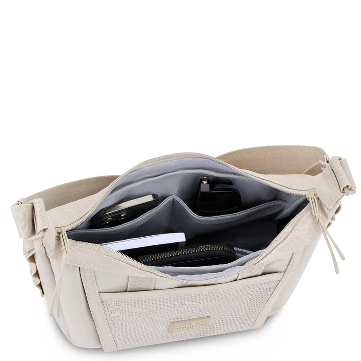 Eine beigefarbene Umhängetasche, deren Oberseite geöffnet ist und in deren Fächern ein Telefon, ein schwarzes Portemonnaie, ein Notizbuch, eine kleine Flasche und andere kleine Accessoires Platz finden.