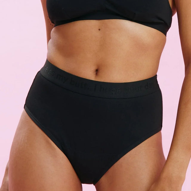 Eine Person, die schwarze Unterwäsche mit hoher Taille und einen passenden schwarzen BH trägt, steht vor einem hellrosa Hintergrund. Das Bild konzentriert sich auf den Oberkörper und die Oberschenkel.