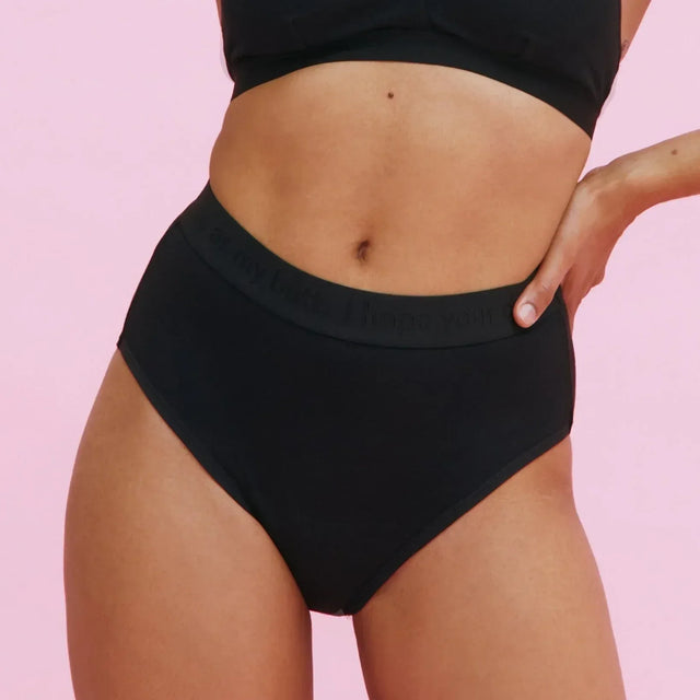 Eine Person in schwarzer Unterwäsche mit hoher Taille und passendem schwarzen Oberteil steht vor einem hellrosa Hintergrund und stützt eine Hand auf die Hüfte.