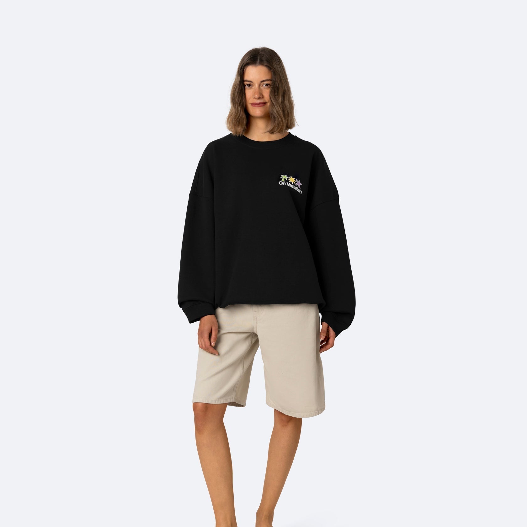 Eine Frau steht mit dem Gesicht nach vorne, trägt ein schwarzes übergroßes Sweatshirt mit einem kleinen Muster auf der Brust, beigefarbene knielange Shorts und schwarze Sandalen vor einem schlichten hellgrauen Hintergrund.