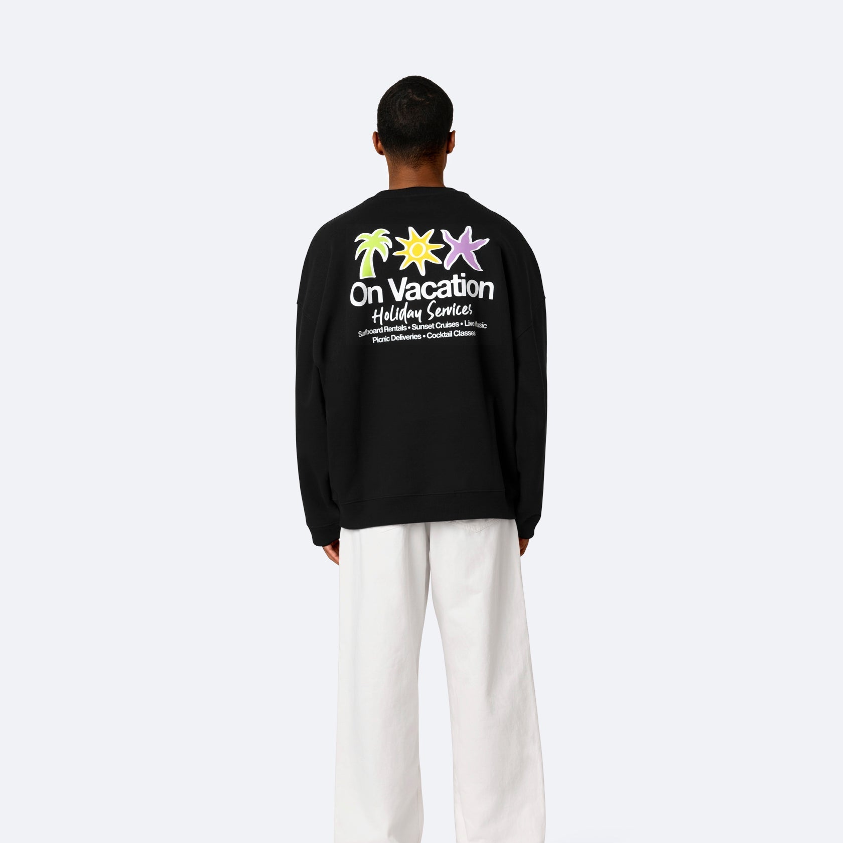 Eine Person steht mit dem Gesicht zur Wand und trägt ein schwarzes Sweatshirt mit bunten Palmen und der Aufschrift On Vacation Holiday Service auf dem Rücken, gepaart mit einer weiten weißen Hose und weißen Schuhen vor einem schlichten Hintergrund.