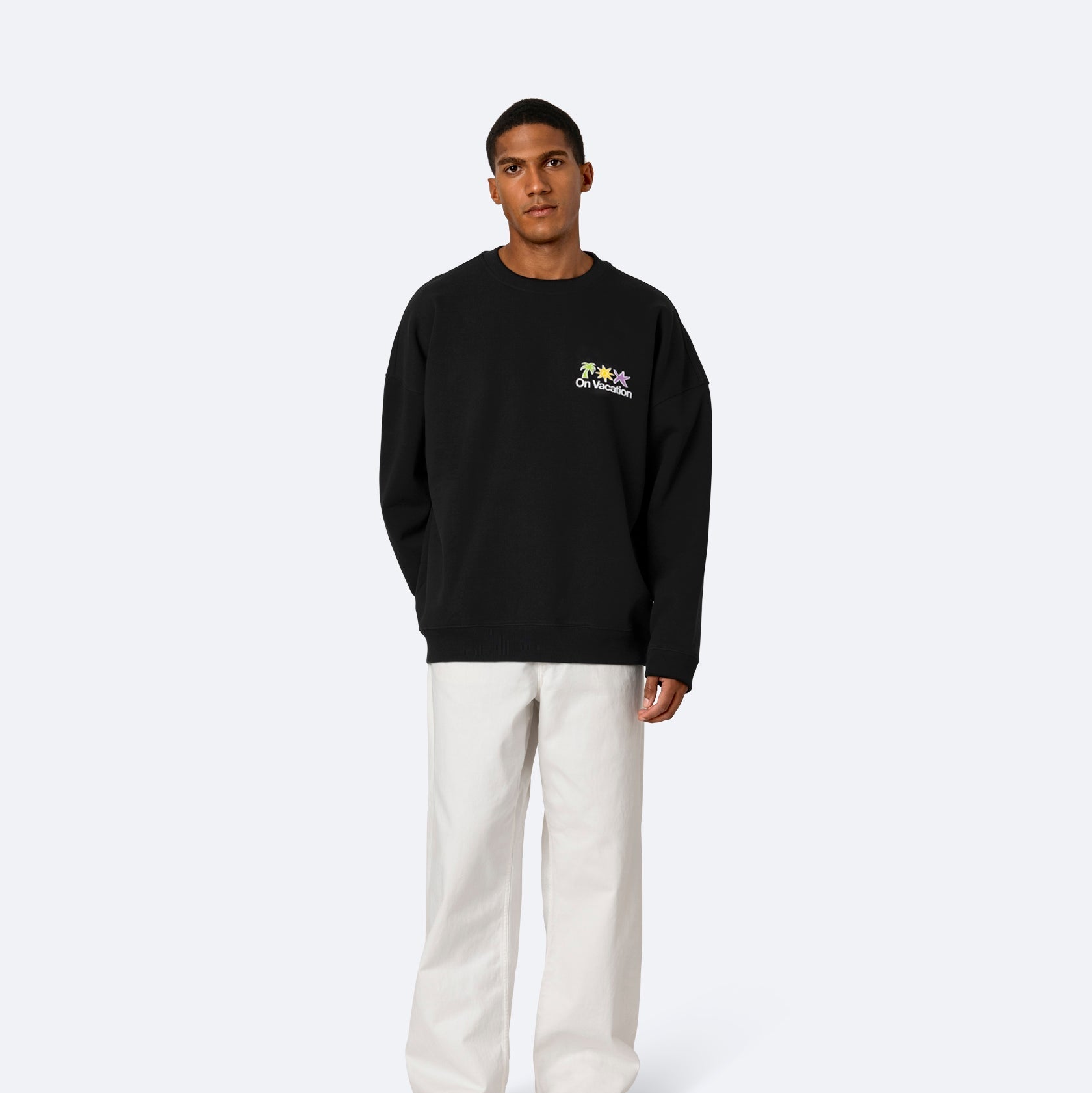 Ein Mann steht mit dem Gesicht nach vorne und trägt ein lockeres schwarzes Sweatshirt mit kleinen bunten Grafiken auf der Brust, eine weiße Hose mit weiten Beinen und weiße Turnschuhe vor einem schlichten hellen Hintergrund.