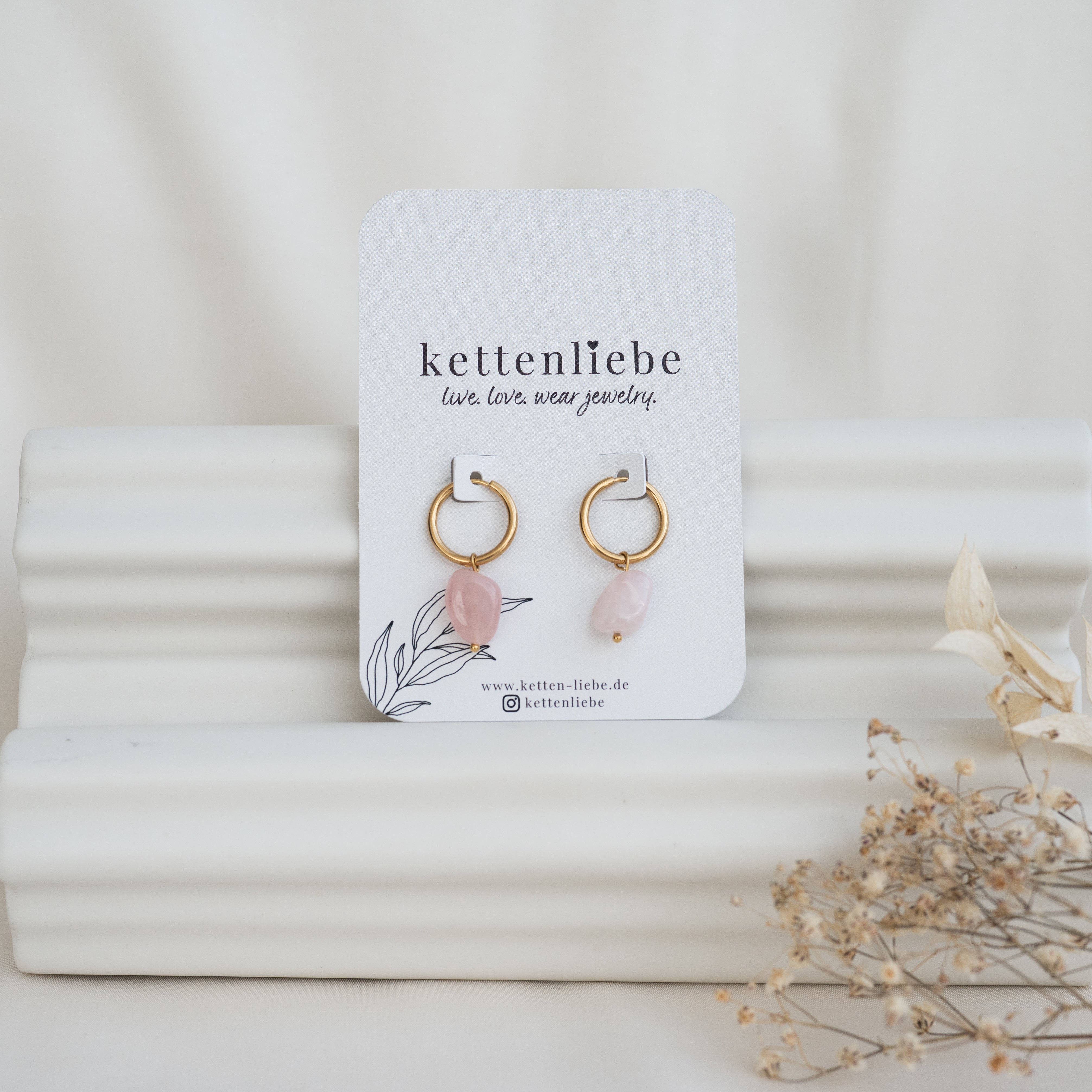 Ein Paar goldene Creolen mit rosafarbenen Steinanhängern auf einer weißen Karte mit der Aufschrift "kettenliebe" vor einem weißen, strukturierten Hintergrund mit getrockneten Blumen auf der rechten Seite.