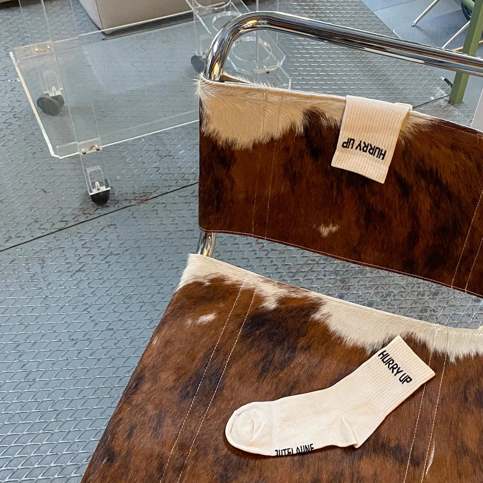 Auf einem braun-weißen Kuhfellstuhl liegen eine einzelne weiße Socke und eine weitere weiße Socke, die über die Rückenlehne drapiert ist. Auf beiden Socken ist der Text HURRY UP aufgedruckt. Der Hintergrund besteht aus einem Metallboden und durchsichtigen Möbeln.