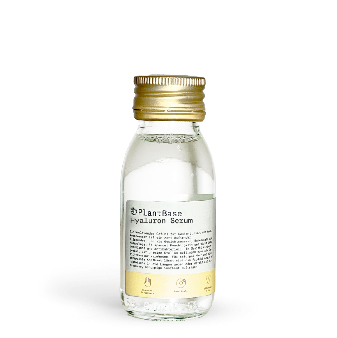 Eine Klarglasflasche mit einem goldenen Verschluss, die PlantBase Hyaluron Serum enthält. Die Flasche ist mit einem weiß-beigen Etikett mit Text und Symbolen versehen, die Details zum Hautpflegeprodukt angeben.