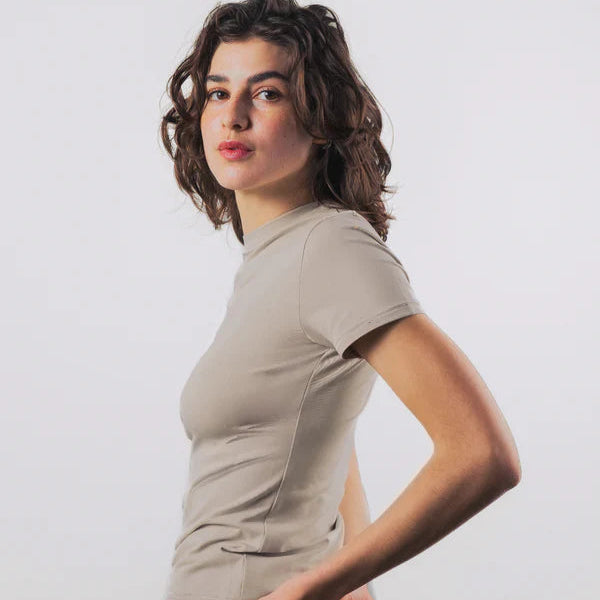 Eine junge Frau mit gewelltem braunem Haar, die ein kurzärmeliges beigefarbenes T-Shirt trägt, steht vor einem schlichten hellen Hintergrund und schaut mit neutralem Blick leicht zur Seite.