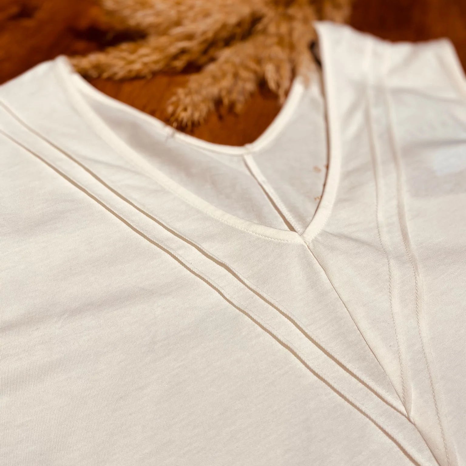 Nahaufnahme von zwei weißen T-Shirts mit V-Ausschnitt, die auf einer hölzernen Oberfläche liegen, mit getrocknetem beigem Pampasgras im Hintergrund. Das Bild hebt die Textur und die Nähte des Stoffes hervor.