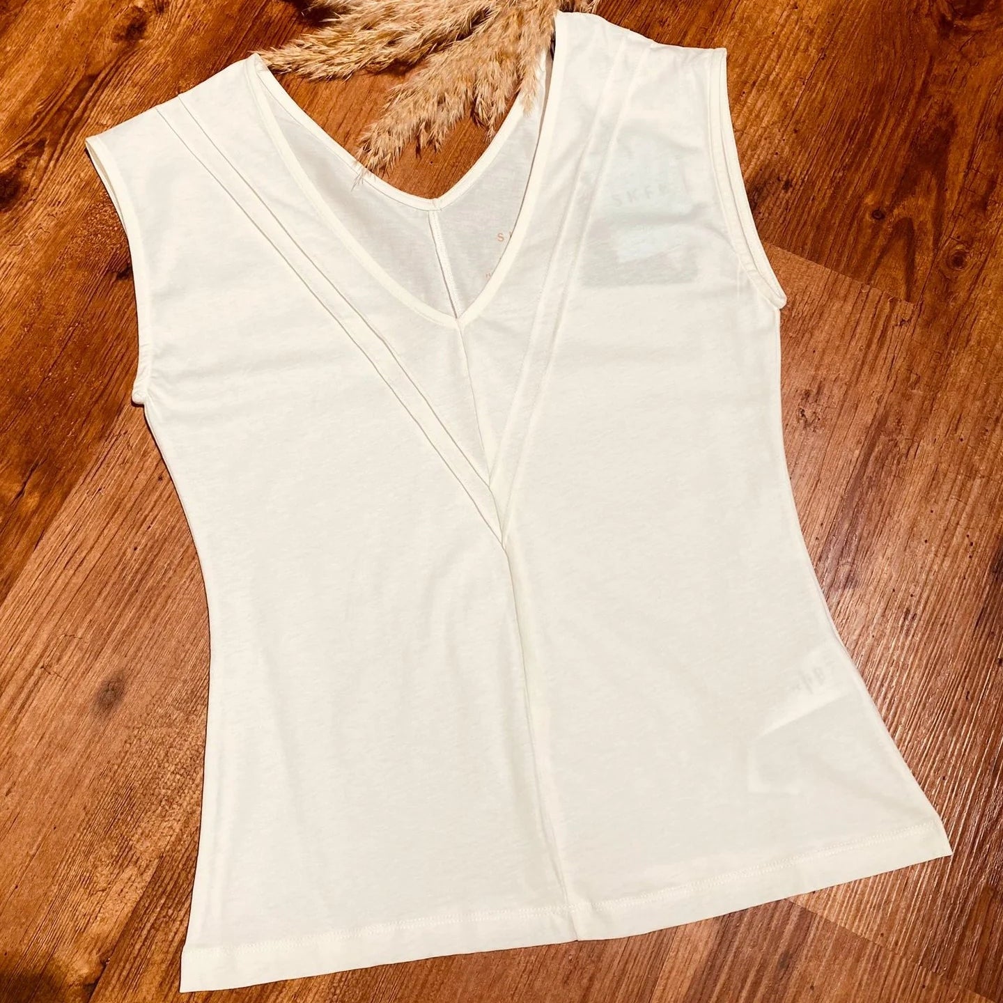 Ein weißes, ärmelloses Oberteil mit V-Ausschnitt und dezenten Nähten liegt flach auf einem Holzboden neben einem Bündel getrockneten Pampasgrases.