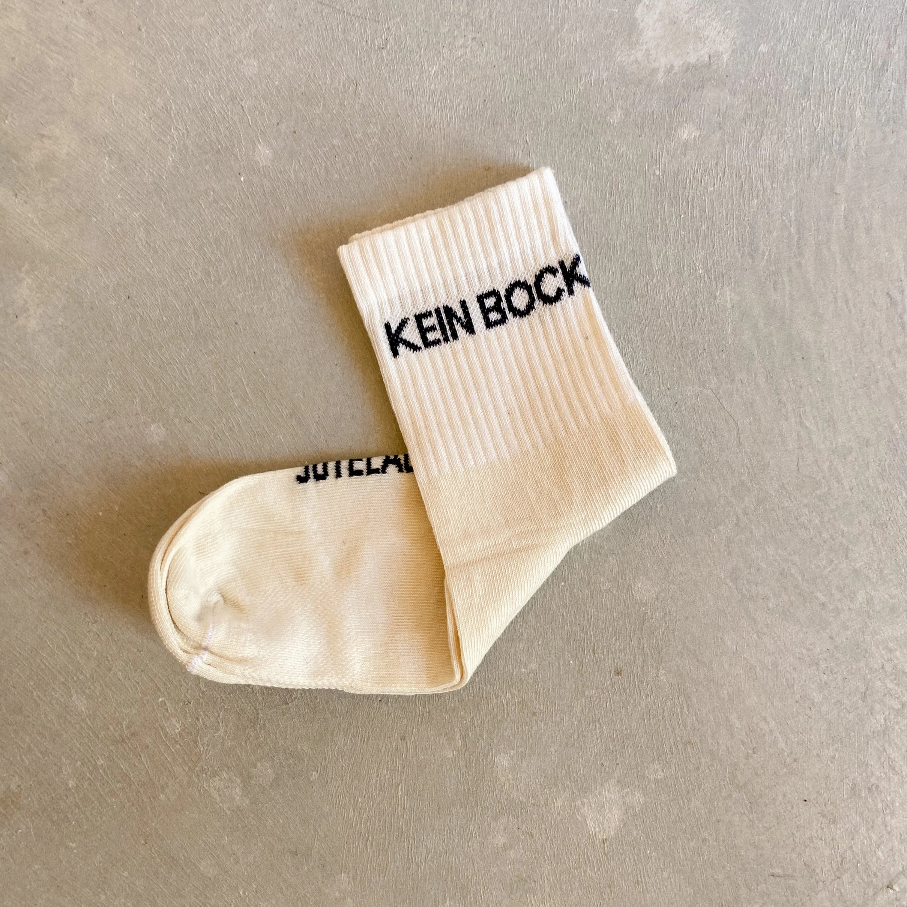 Eine einzelne helle Socke liegt auf einer strukturierten, grauen Oberfläche. Oben auf der Socke steht in schwarzer Schrift KEIN BOCK.