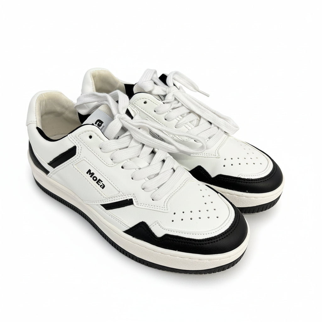 Ein Paar weiße Sneakers mit schwarzen Akzenten und MoEa-Branding an der Seite, dargestellt auf weißem Hintergrund. Die Schuhe haben weiße Schnürsenkel und perforierte Details an den Zehen.
