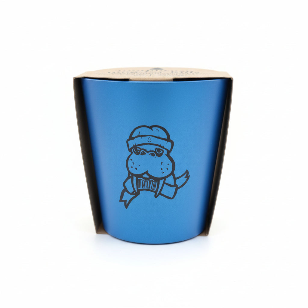 Eine blaue Tasse mit einem Cartoon-Nilpferd, das eine Brille, einen Schal und einen Hut trägt, mit schwarzen Akzenten an den Seiten, vor einem weißen Hintergrund.