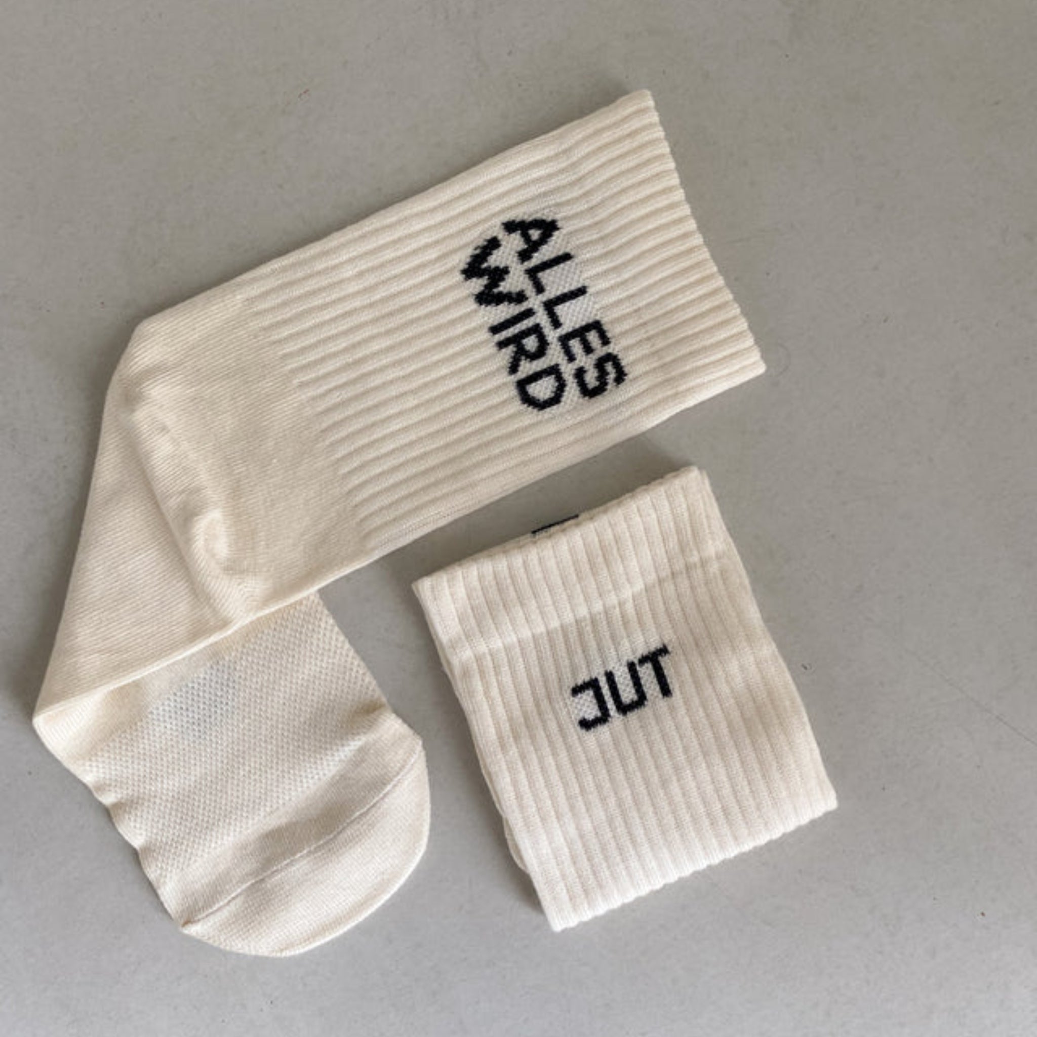 Ein Paar cremefarbener gerippter Socken auf einer hellen Oberfläche. Eine Socke trägt die Aufschrift ALLES WIRD und die andere die Aufschrift GUT, die zusammen den deutschen Satz ALLES WIRD GUT bilden.