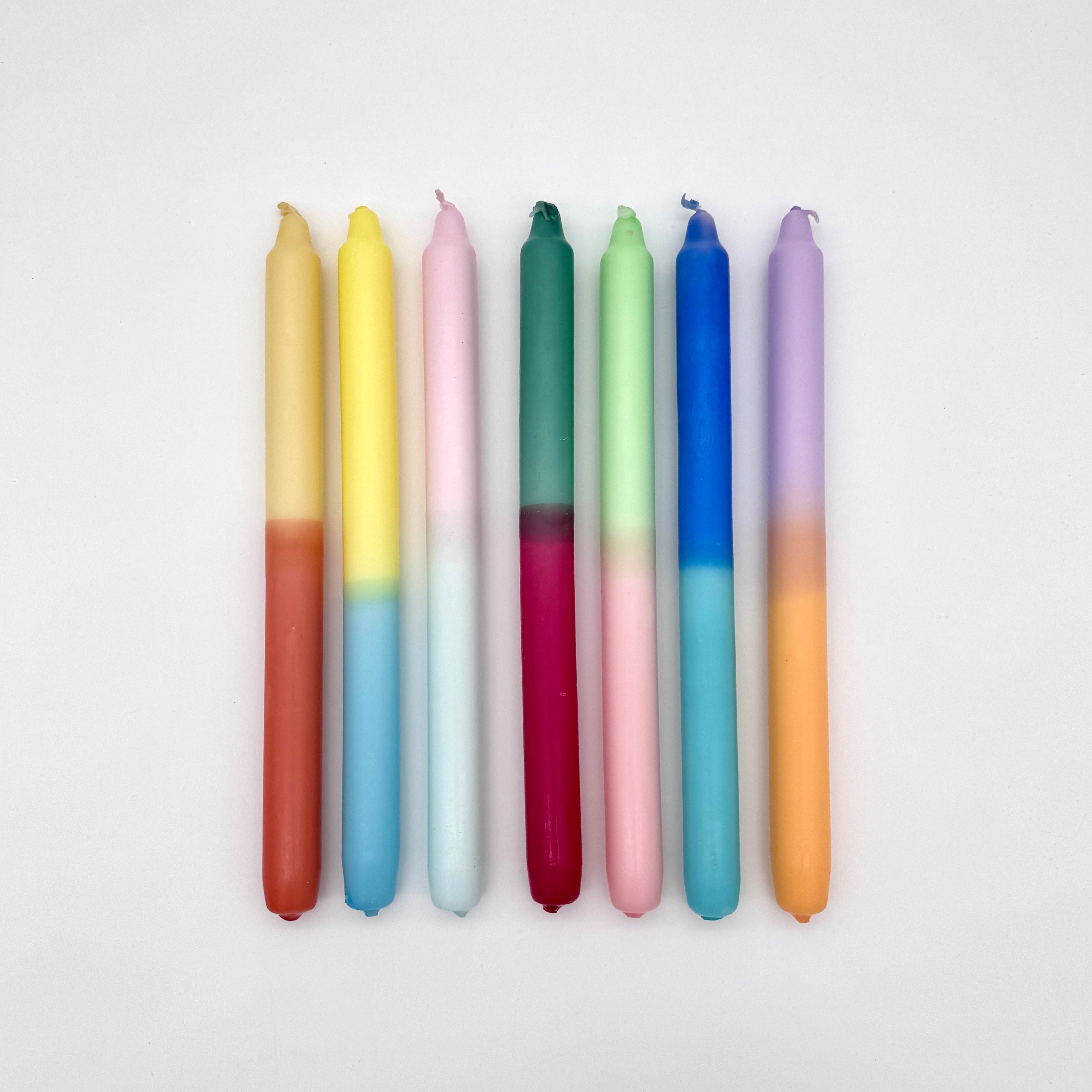 Sieben bunte, zweifarbige Spitzkerzen sind vertikal nebeneinander auf einem weißen Hintergrund angeordnet. Jede Kerze hat zwei verschiedene Farben, die sich in der Mitte vermischen.