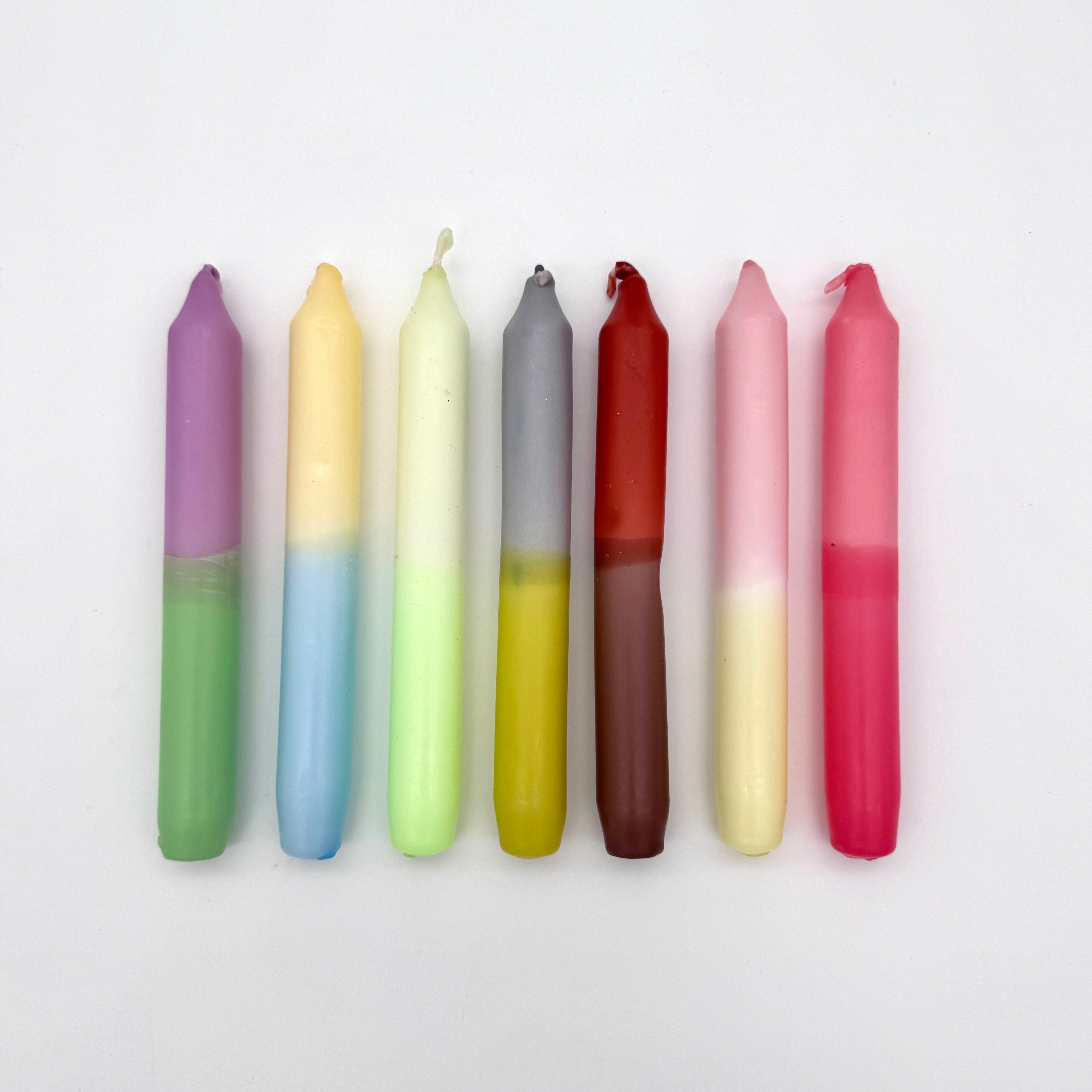 Sieben bunte, vertikal gestreifte Kerzen in Pastell- und kräftigen Tönen sind in einer Reihe auf weißem Hintergrund angeordnet. Jede Kerze hat zwei unterschiedliche Farbabschnitte.