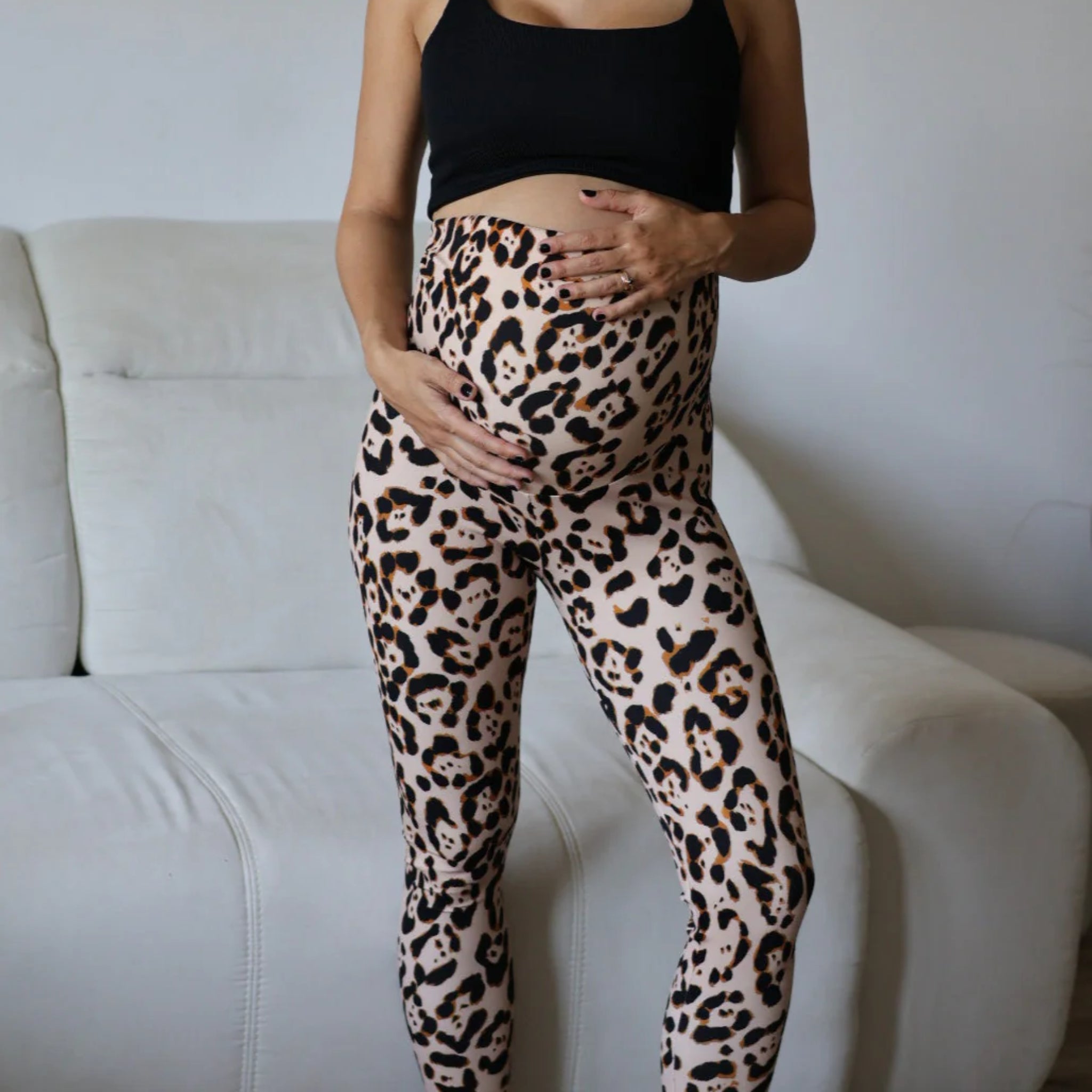 Eine schwangere Frau, die ein schwarzes Crop-Top und Leggings mit Leopardenmuster trägt, steht im Haus vor einem hellen Sofa und streichelt mit beiden Händen sanft ihren Bauch.