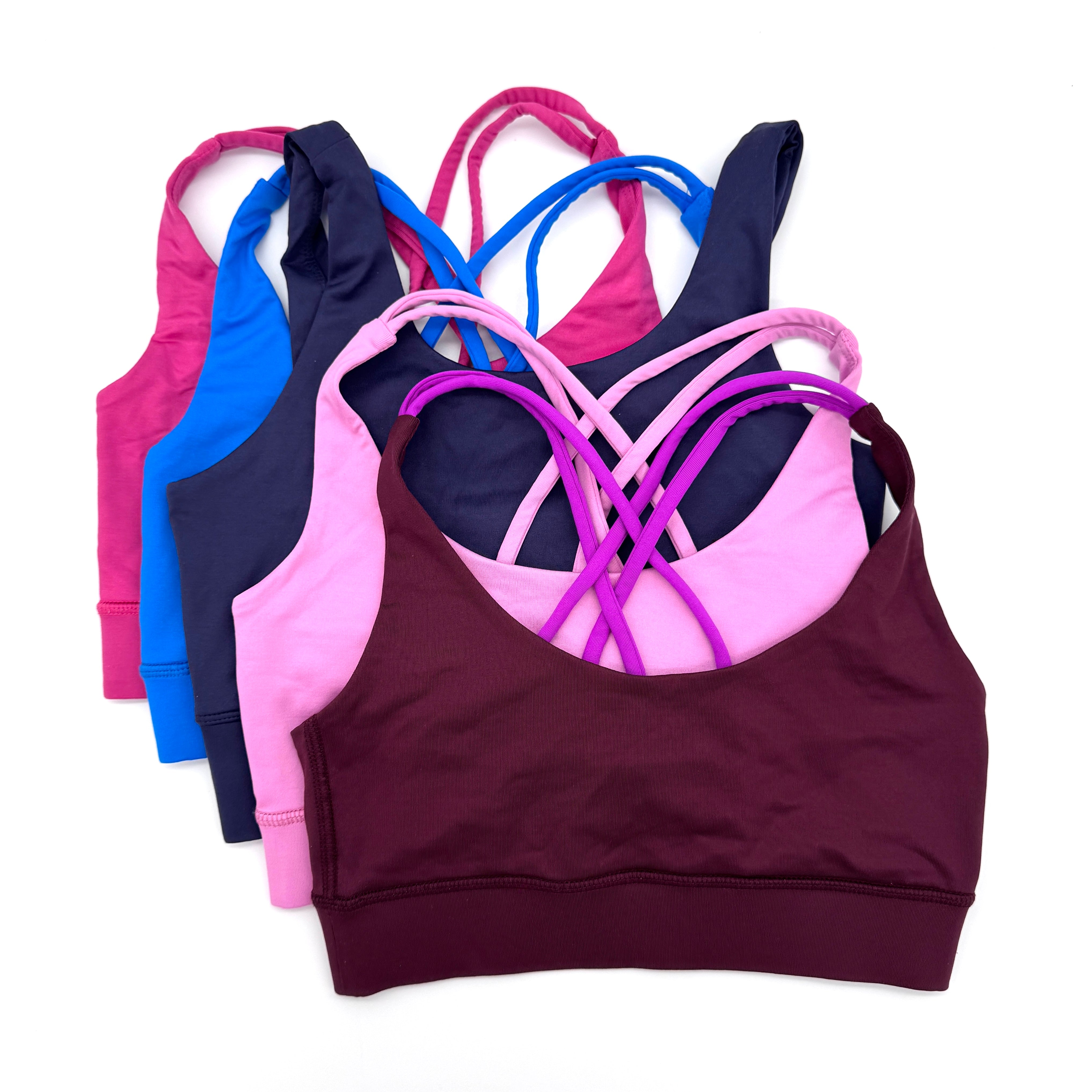 Fünf Sport-BHs in verschiedenen Farben (weinrot, hellrosa, marineblau, blau und rosa) sind in einem überlappenden Stapel auf weißem Hintergrund angeordnet und zeigen gekreuzte Träger.