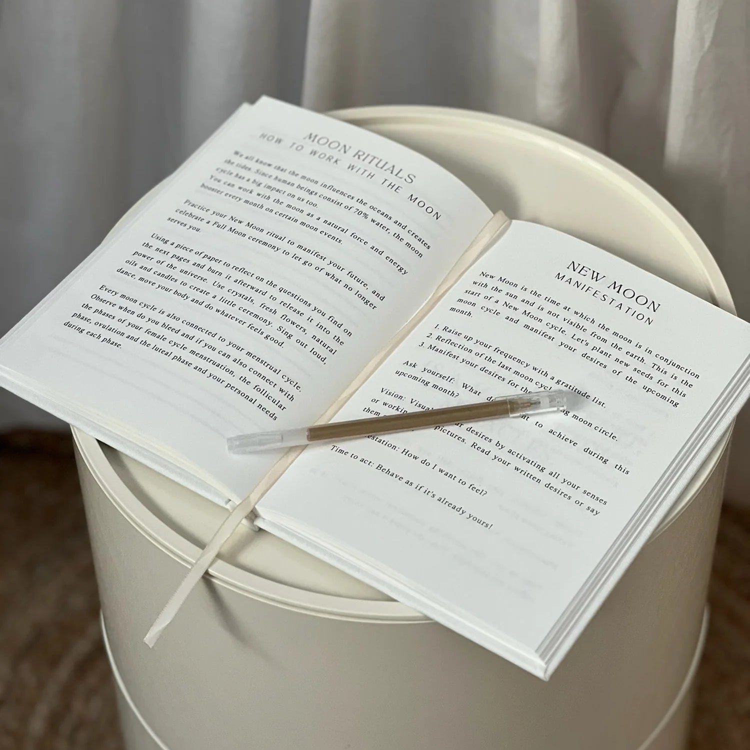Auf einem runden weißen Tisch liegt ein aufgeschlagenes Buch, das Seiten über Mondrituale und Neumond-Affirmationen enthält. Ein beigefarbenes Lesezeichen liegt quer über die Seiten. Im Hintergrund hängen helle Vorhänge.