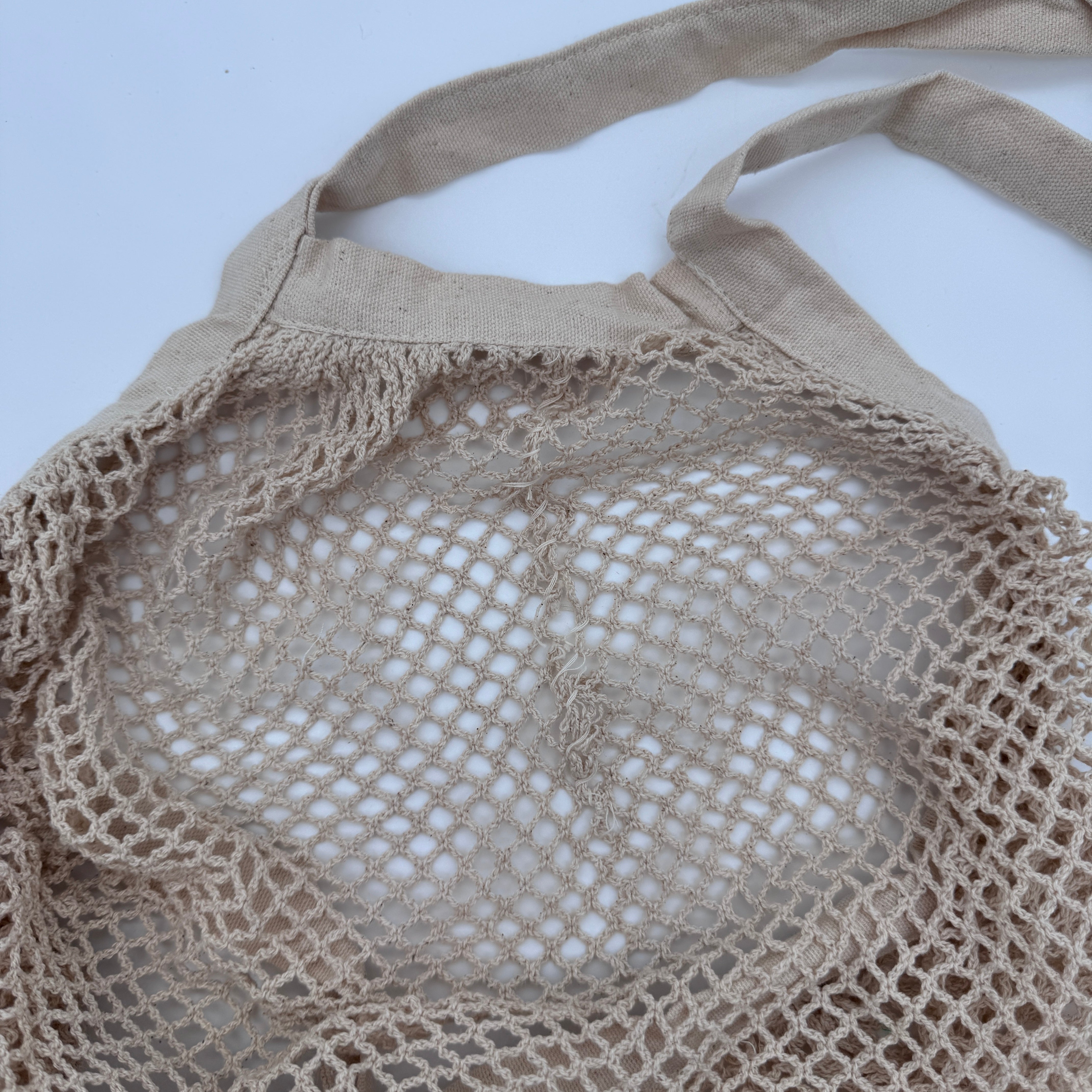 Eine beigefarbene wiederverwendbare Einkaufstasche aus Baumwollgewebe mit Stoffgriffen liegt flach auf einer weißen Fläche.
