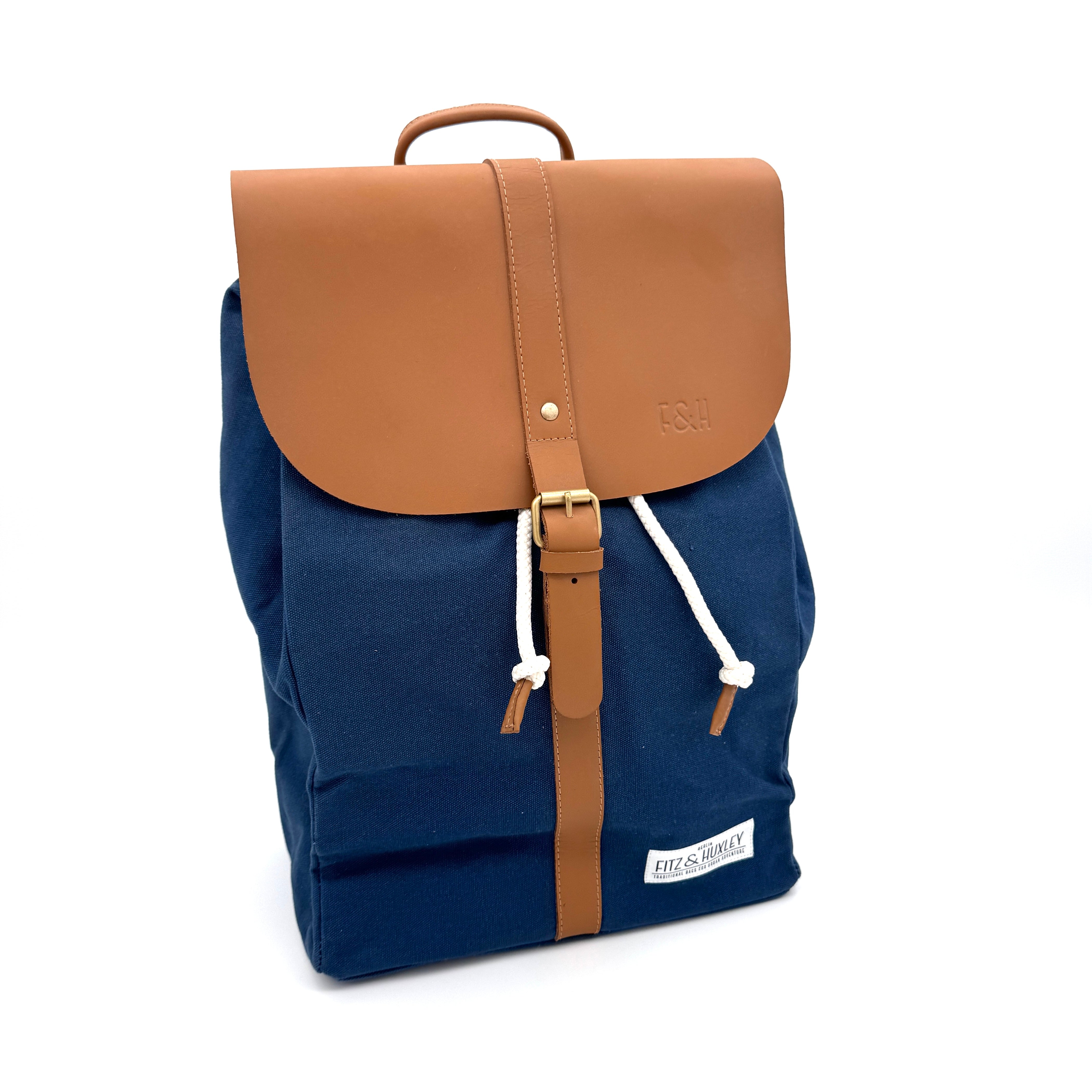 Ein marineblauer Rucksack aus Segeltuch mit einer hellbraunen Lederklappe und einem Griff, weißen Kordelzügen und einem hellbraunen Schnallenriemen. Ein kleines weißes Patch-Etikett mit der Aufschrift Fitz & Huxley auf der unteren Vorderseite.