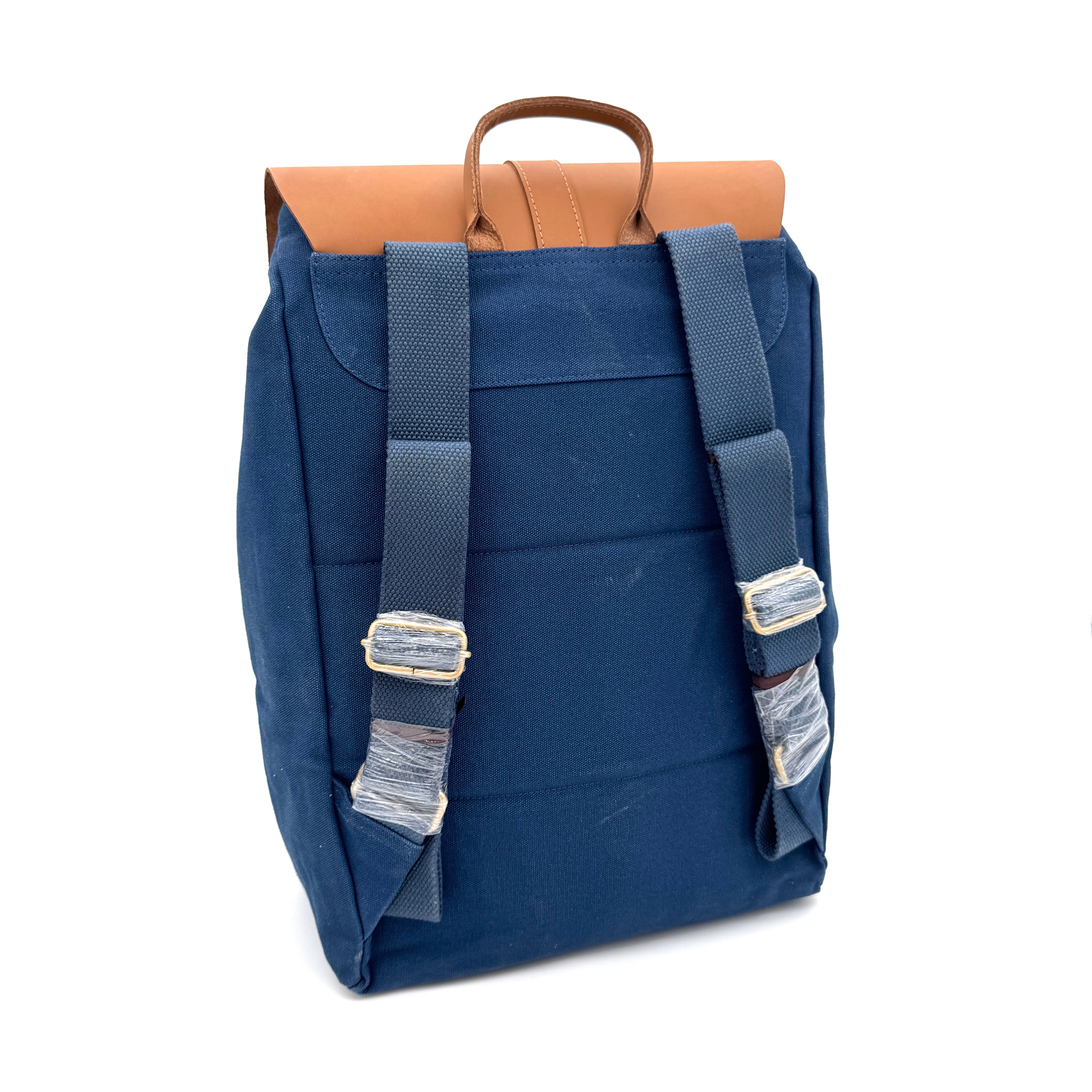 Ein blauer Rucksack aus Segeltuch mit verstellbaren blauen Schulterriemen, silbernen Schnallen, die teilweise mit Plastik umwickelt sind, und einem braunen Ledergriff, dargestellt vor einem weißen Hintergrund.