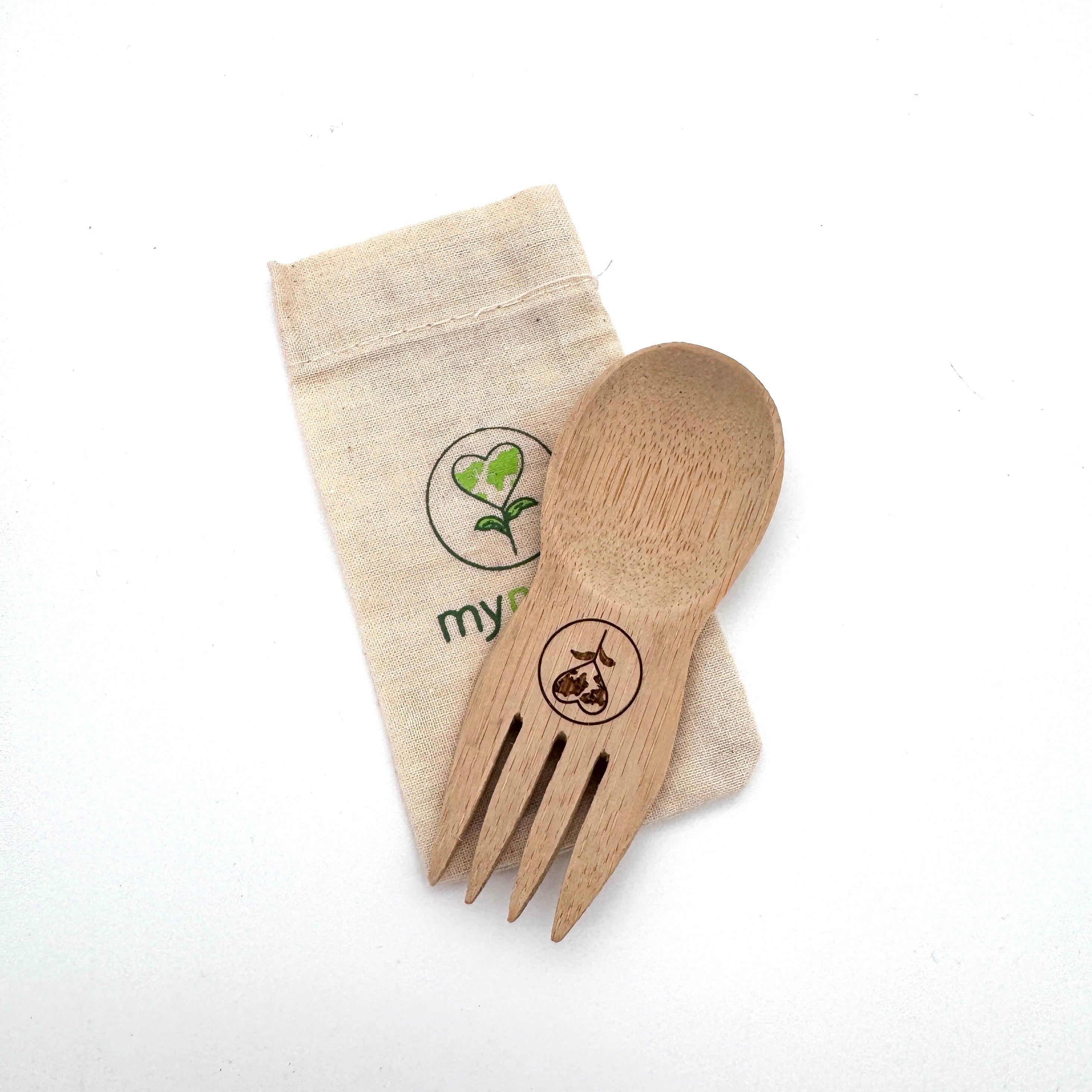 Ein Holzspieß mit eingraviertem Motiv ruht auf einem kleinen beigen Stoffbeutel. Der Beutel hat ein grünes Pflanzenlogo und das Wort my ist teilweise sichtbar.