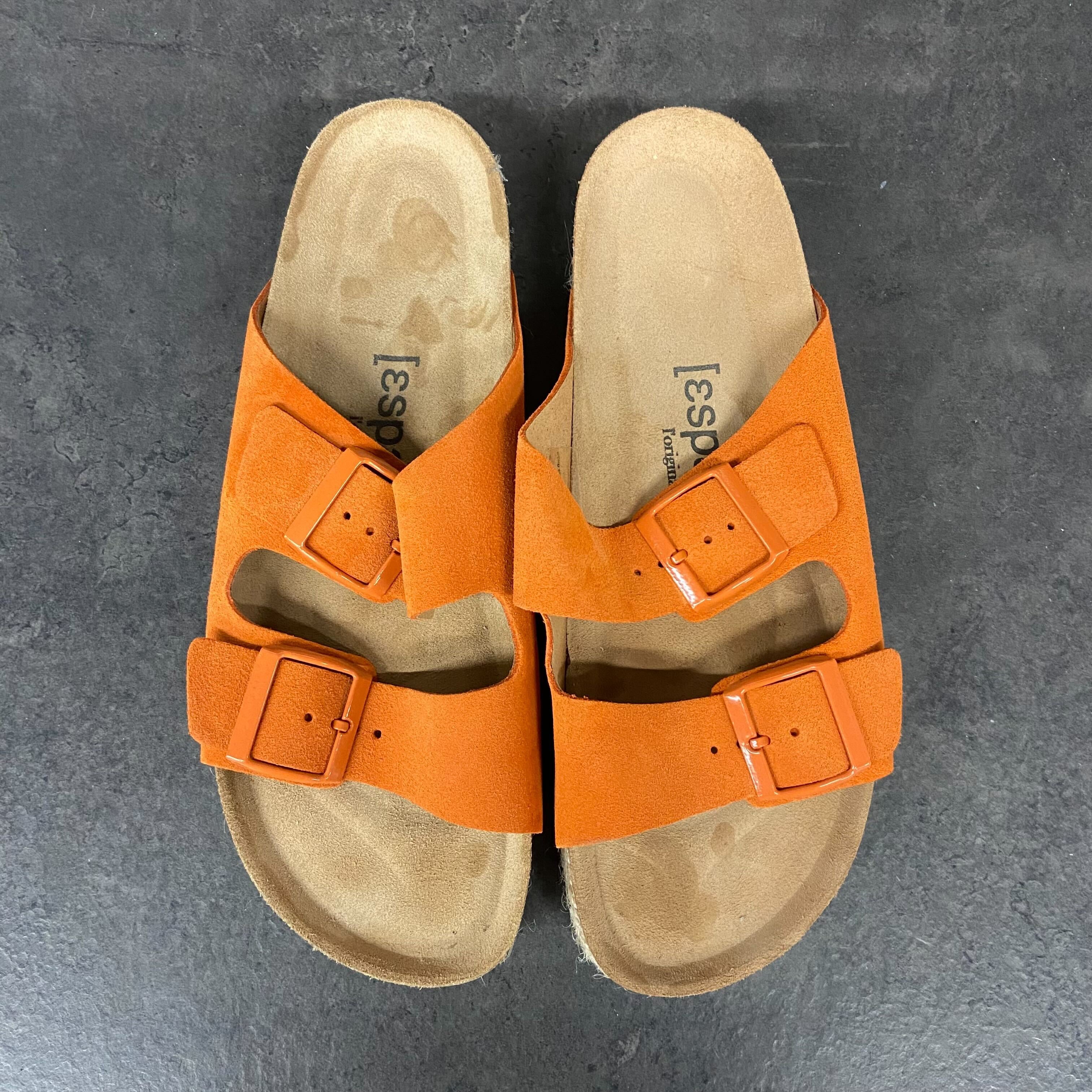 Orange Sandalen mit zwei Schnallen auf dunklem Boden.
