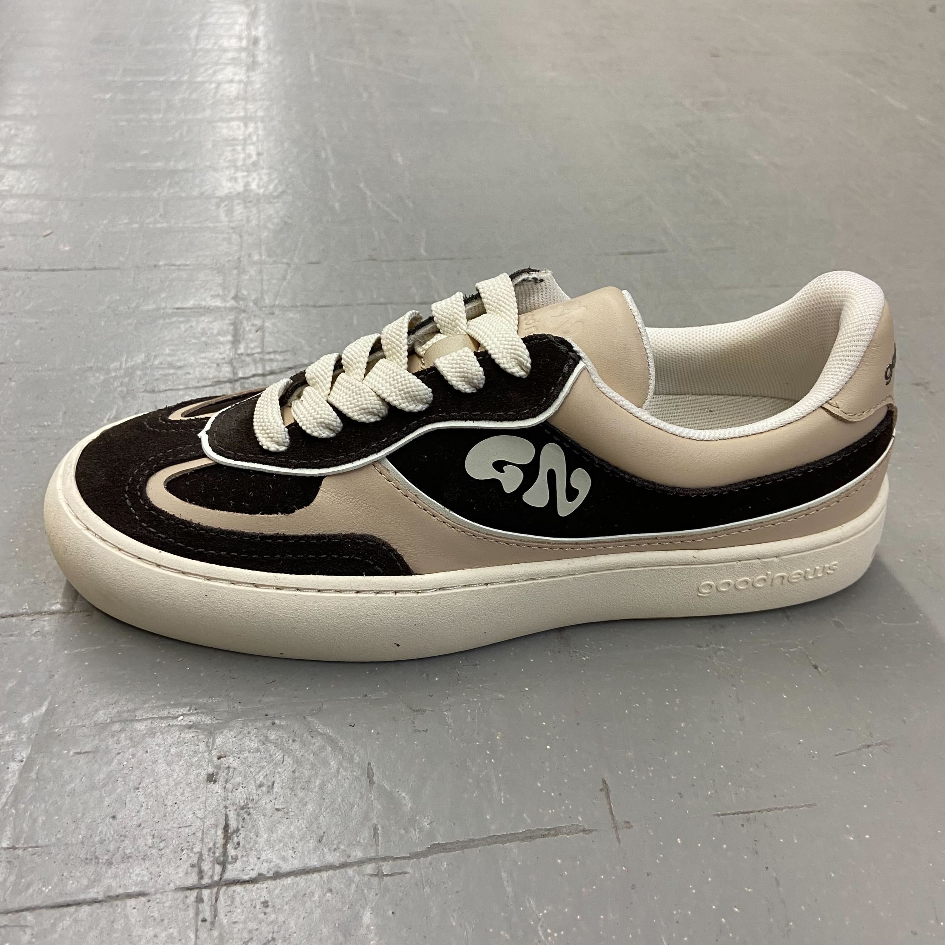 Schwarz-beiger Sneaker mit weißer Sohle und Schnürsenkeln auf grauem Boden.